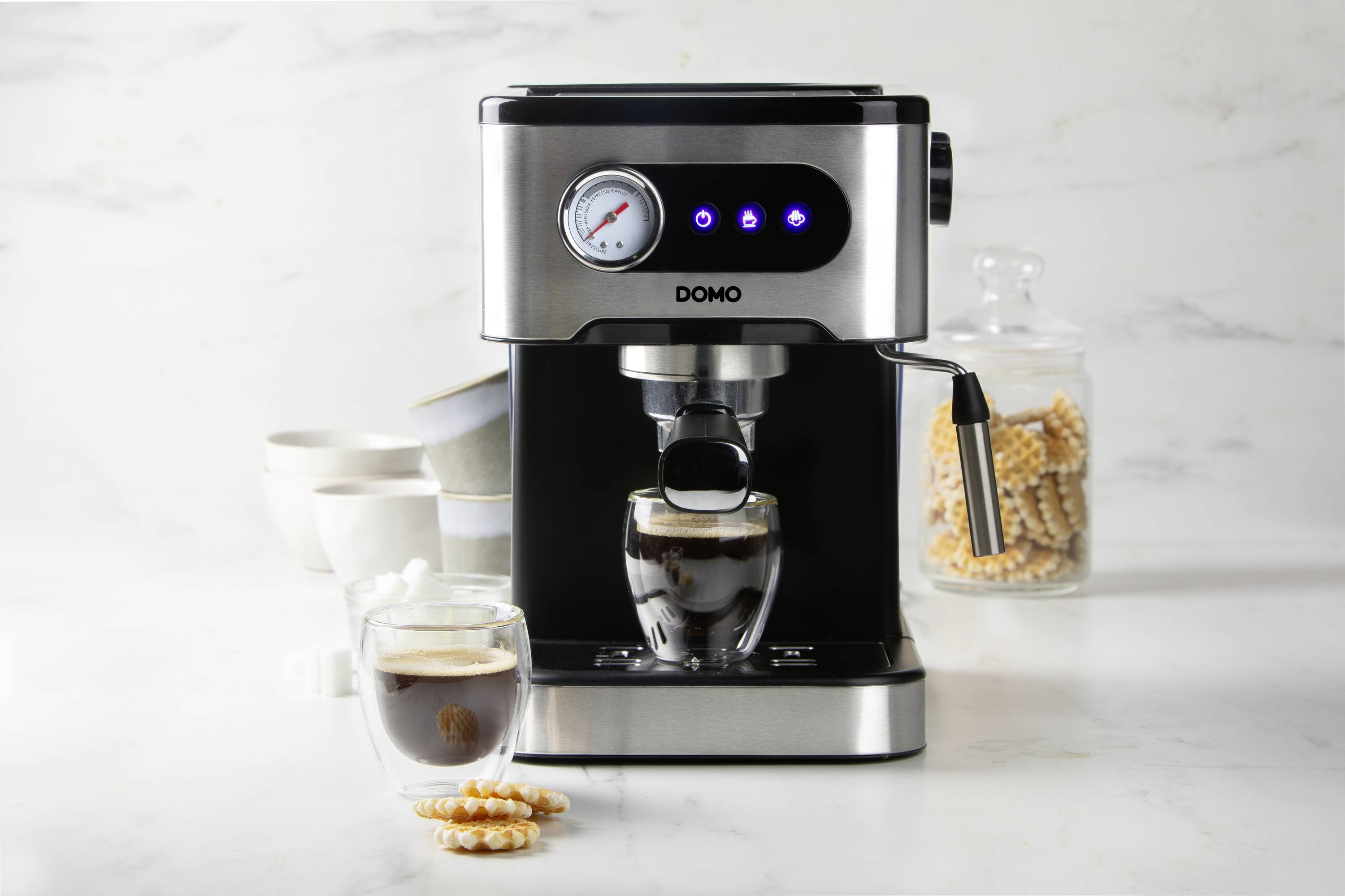 Machine à espresso en acier inoxydable versant un espresso frais dans un verre. À côté, des tasses et un verre avec des biscuits sont posés sur une table.