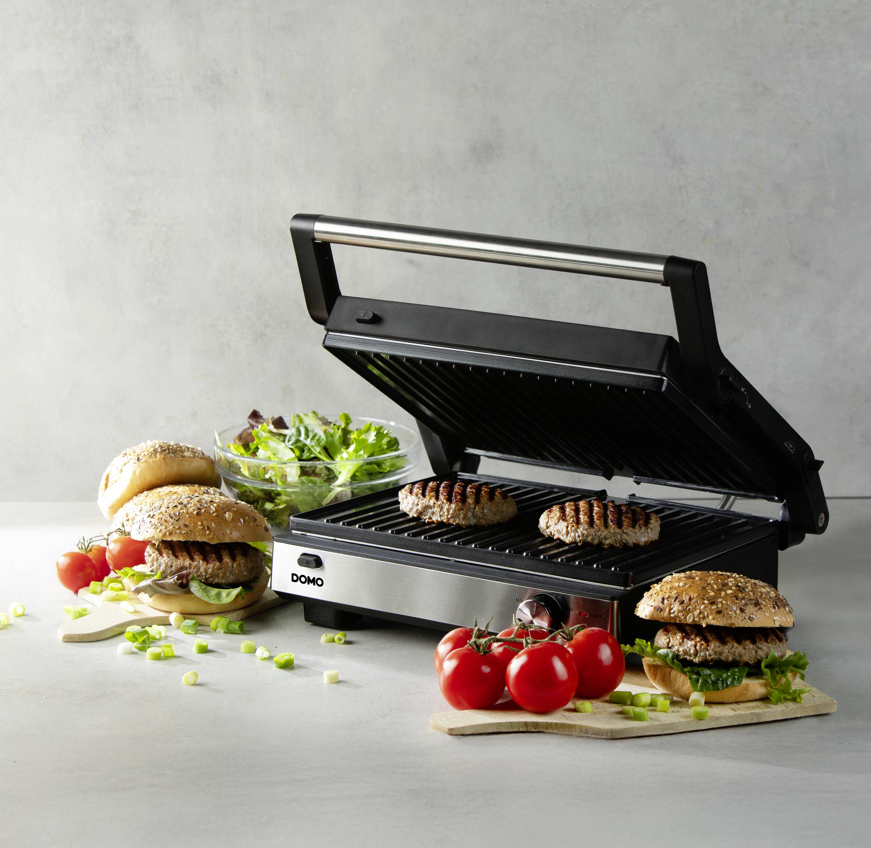 Un grill à contact ouvert avec deux steaks de hamburger. À côté, des hamburgers préparés sont disposés avec de la salade et des tomates en garniture.