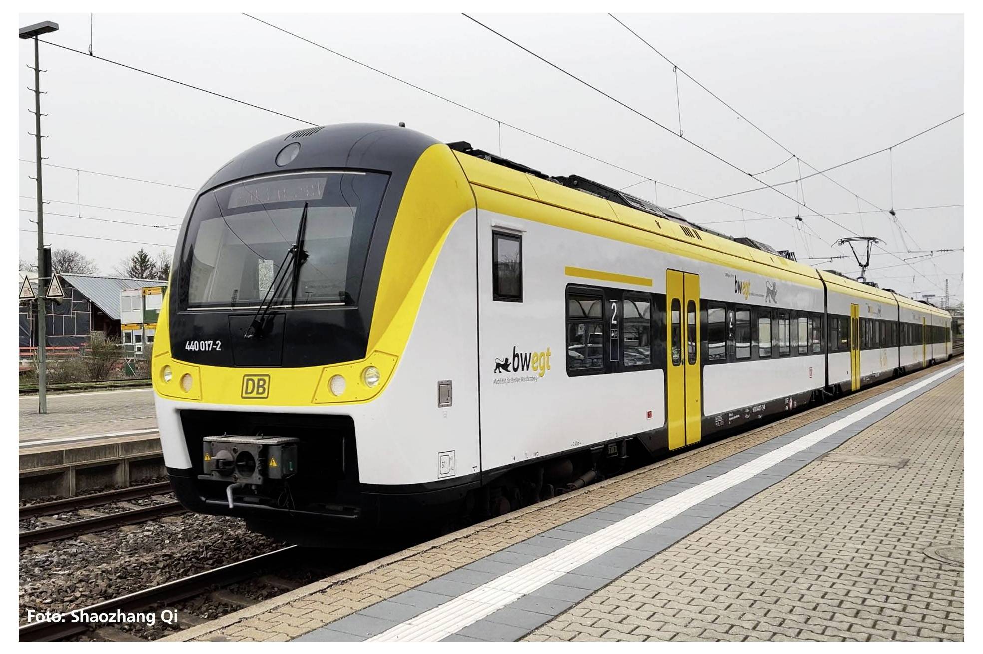 Un train régional jaune portant l'inscription 'bwegt' est stationné sur le quai. Des voies ferrées et des bâtiments sont visibles en arrière-plan.