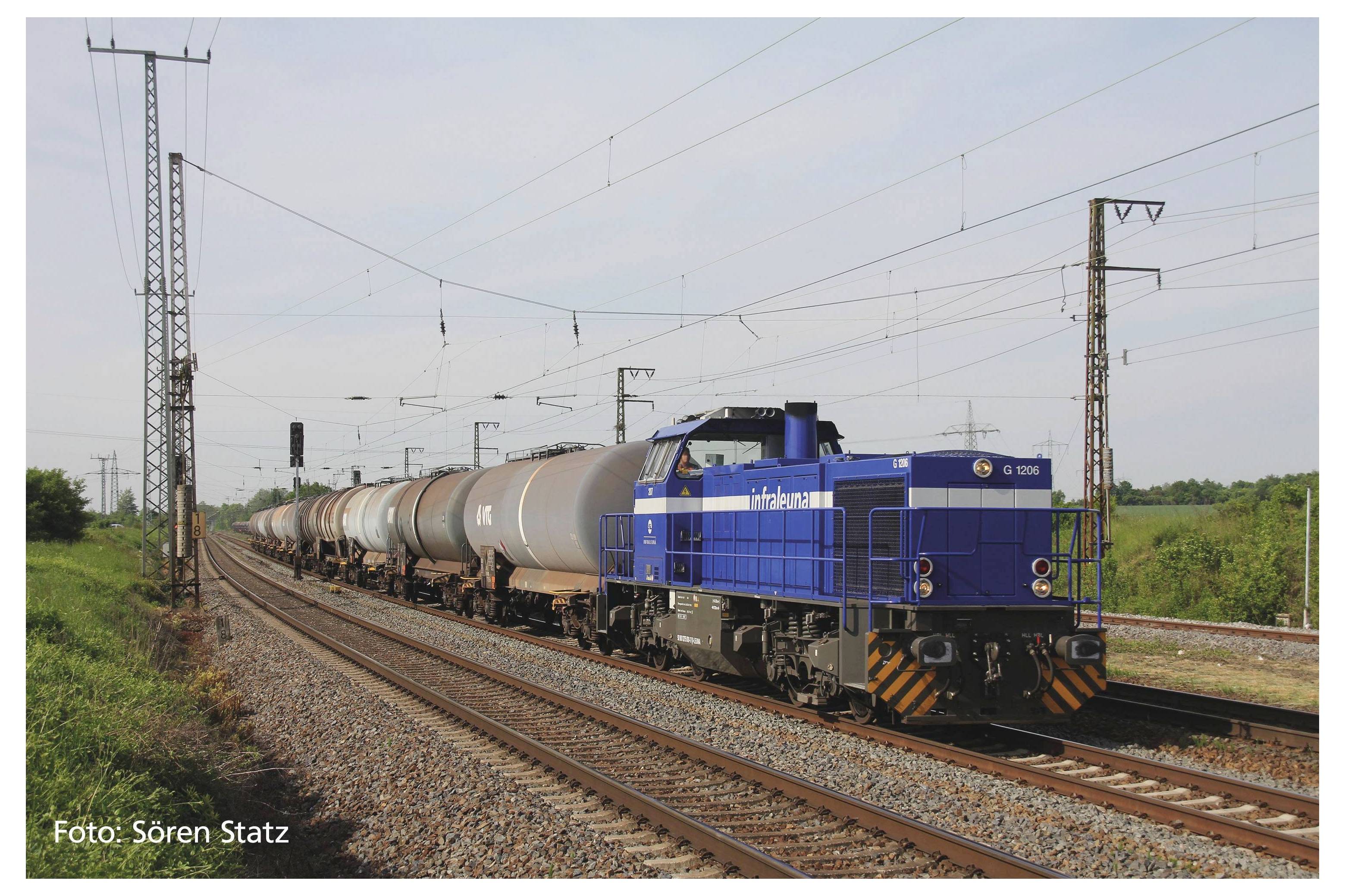 Un train de marchandises bleu transporte plusieurs wagons-citernes sur une voie ferrée à double voie, entouré de paysage verdoyant et de pylônes électriques.