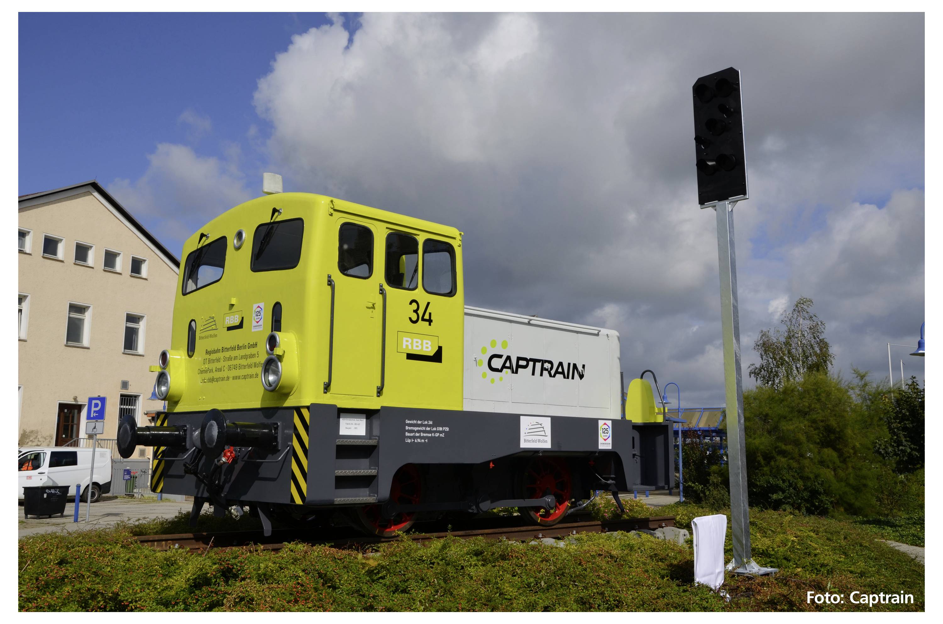 Une petite locomotive jaune portant l'inscription 'Captrain' est stationnée sur un tronçon de voie ferrée en extérieur. Un bâtiment est visible en arrière-plan.