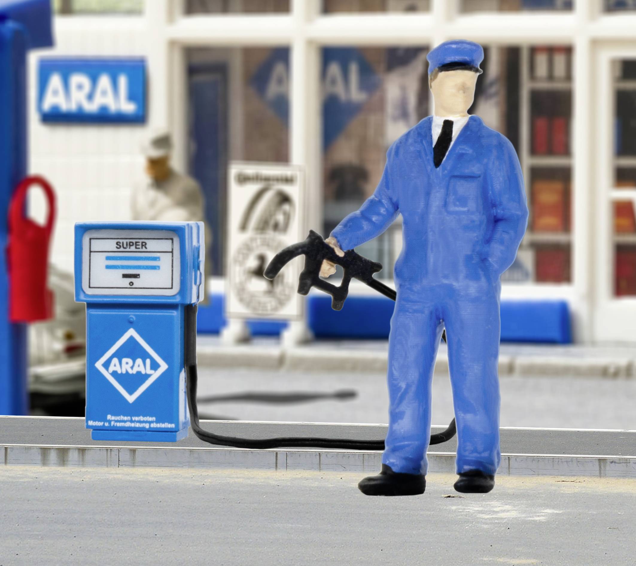 Un modèle d'une station-service avec une figurine en uniforme bleu tenant un pistolet de pompe à essence. Un panneau ARAL est visible en arrière-plan.