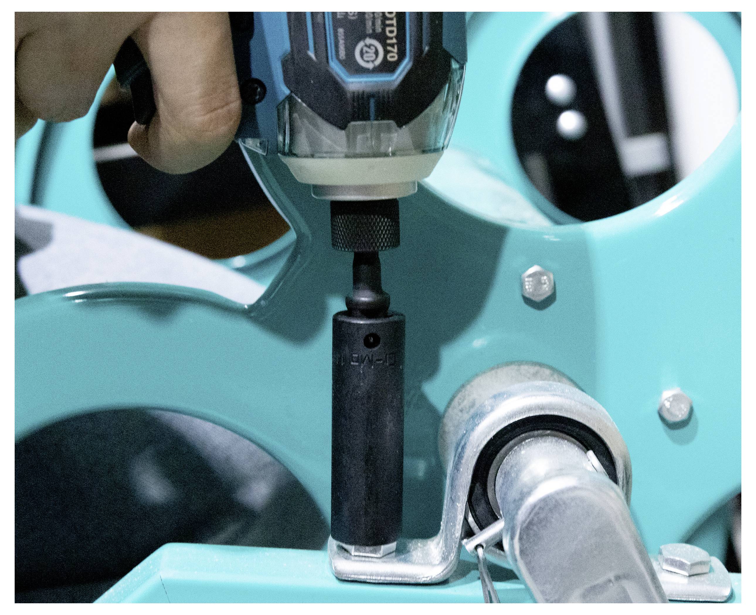 Une personne utilise une perceuse avec un embout de socket pour ajuster un composant métallique d'une machine turquoise, en resserrant un boulon.