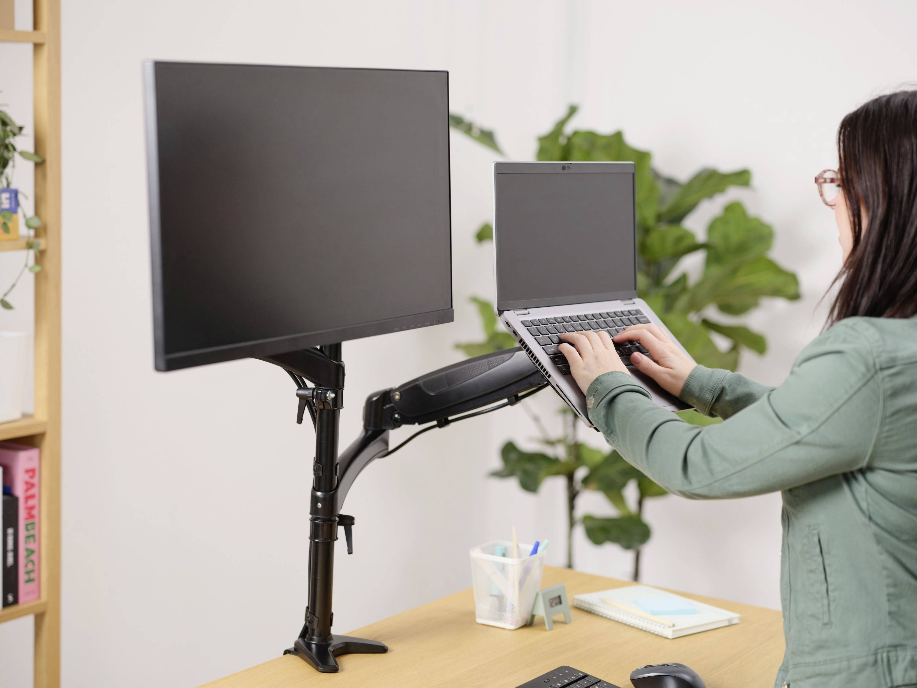 Une personne est debout à un bureau, utilisant un ordinateur portable et deux écrans sur un support ajustable. Une plante se trouve en arrière-plan.