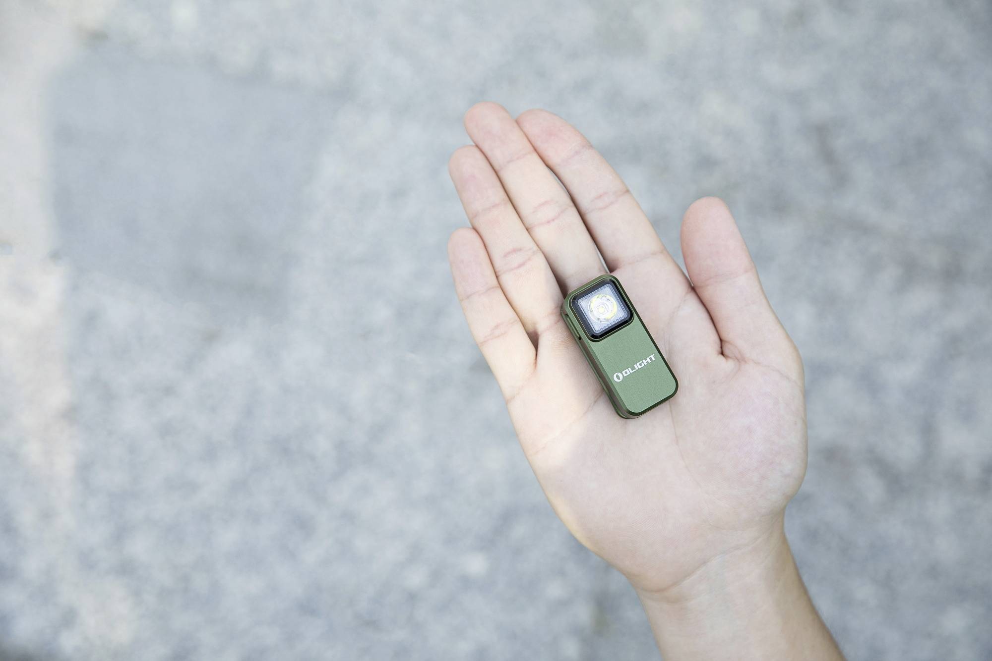 Une personne tient une petite lampe de poche verte allumée dans sa paume. L'arrière-plan est gris et flou.