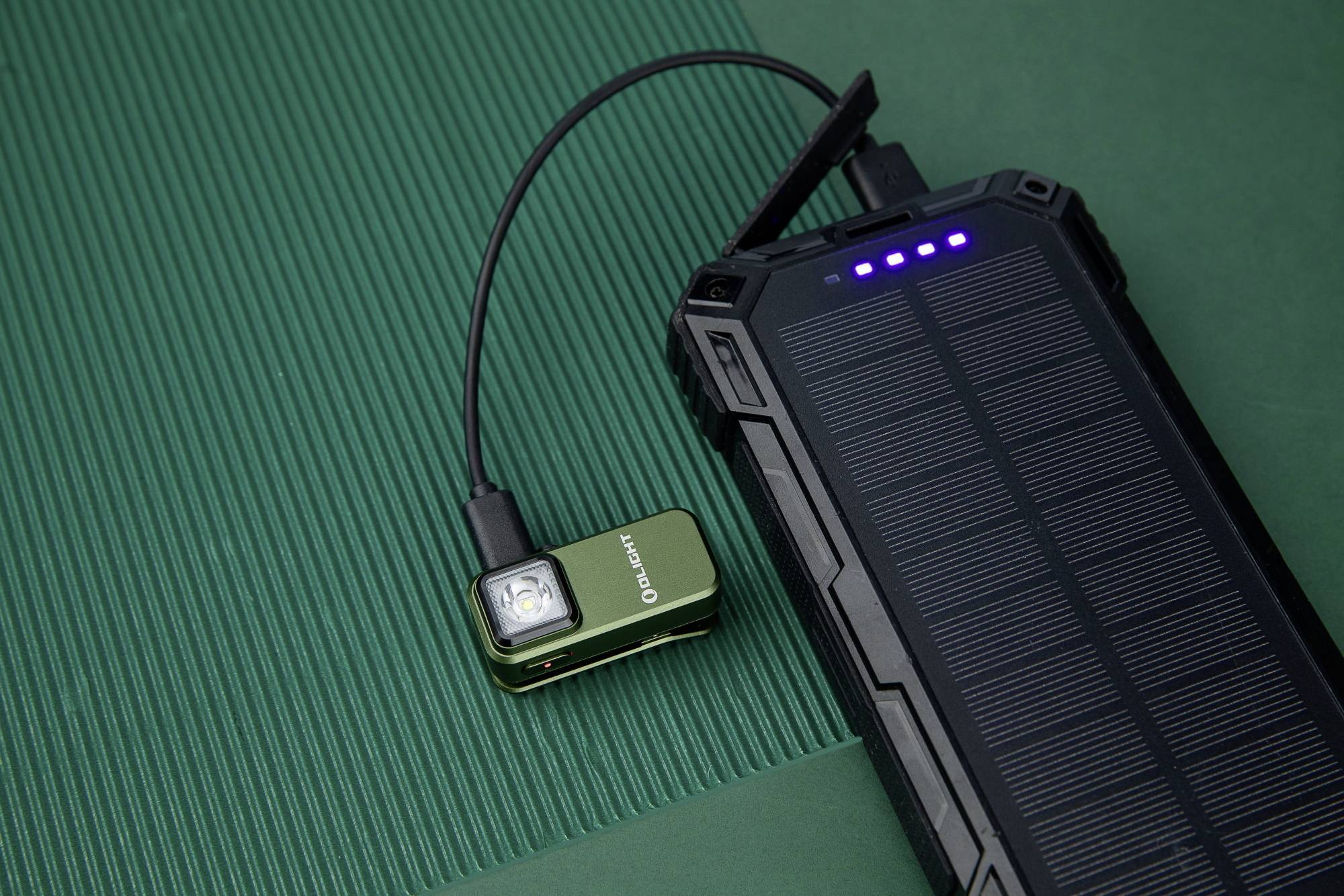 Batterie externe avec module solaire charge une lampe de poche verte et compacte par câble. Les deux appareils sont posés sur une surface verte.