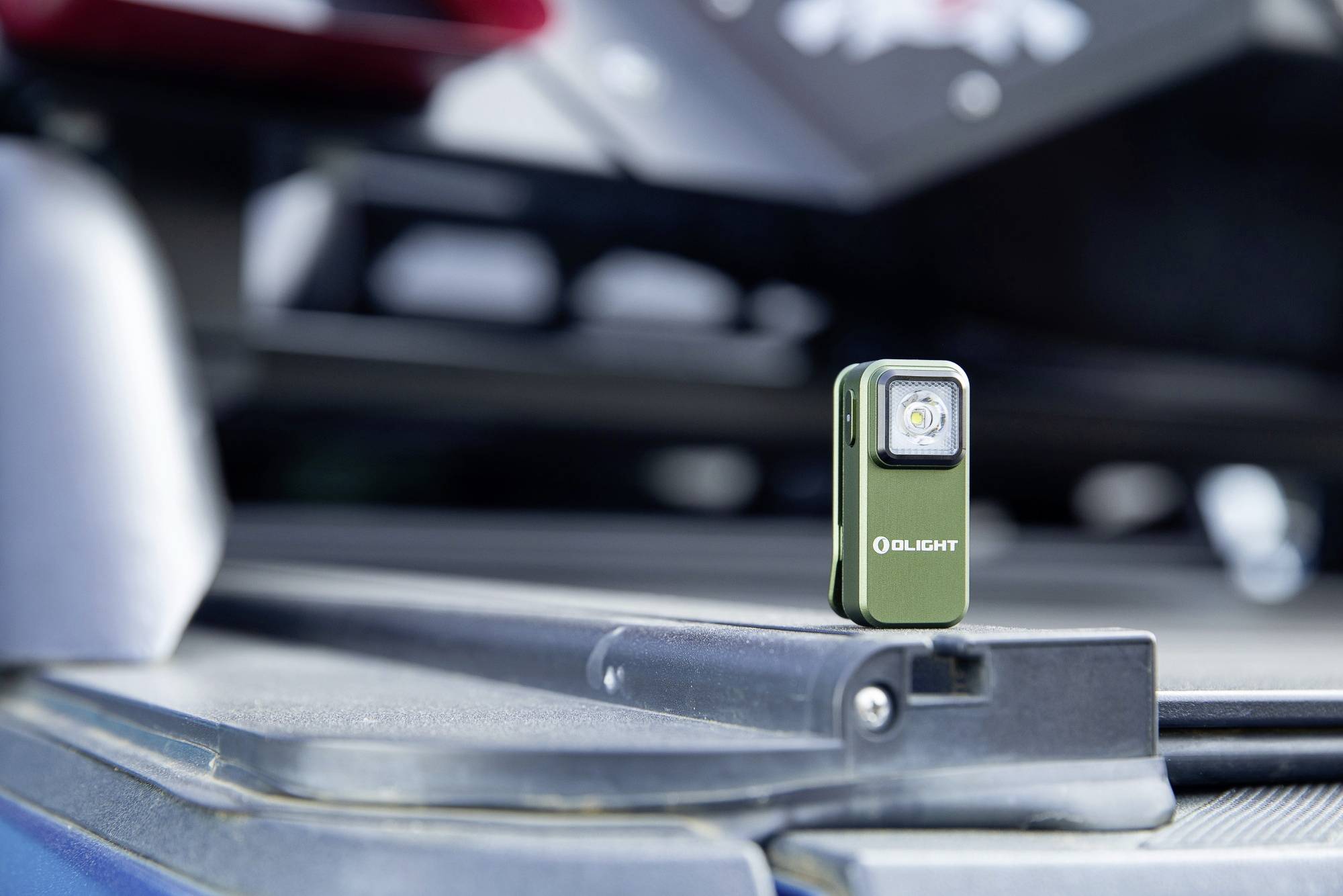 Petit cube vert de lampe de poche avec logo de marque, placé sur une surface lisse, avec des détails de véhicule flous en arrière-plan.