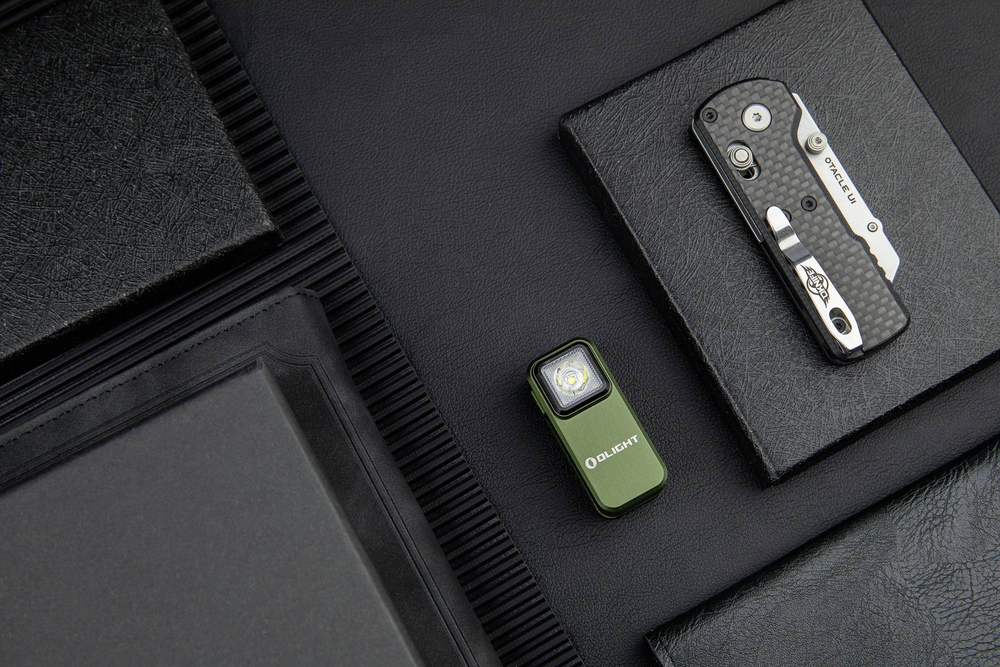Une petite lampe de poche et un couteau pliant sont posés côte à côte sur une surface en cuir noir. Les deux objets sont compacts et conçus de manière fonctionnelle.