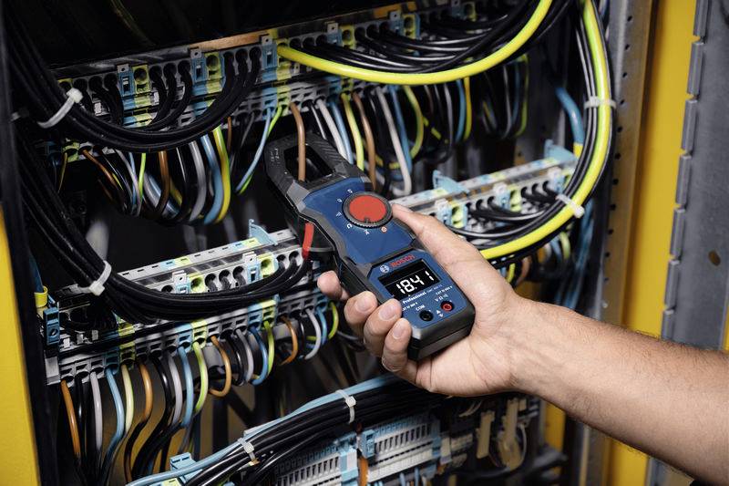 Une main tient un appareil de mesure numérique et mesure le courant dans un armoire électrique contenant plusieurs câbles et connexions.