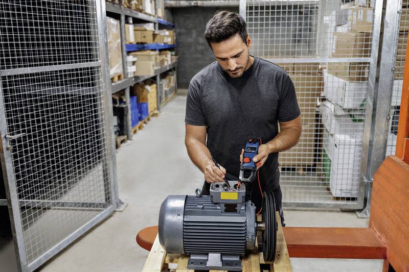 Un homme vérifie un grand moteur à l'aide d'un appareil de mesure dans un local d'entreposage. Des étagères chargées de cartons sont visibles en arrière-plan.