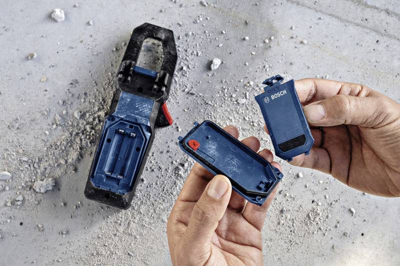 Une personne tient un outil Bosch bleu ouvert sur un chantier de construction, entourée de poussière et de décombres.