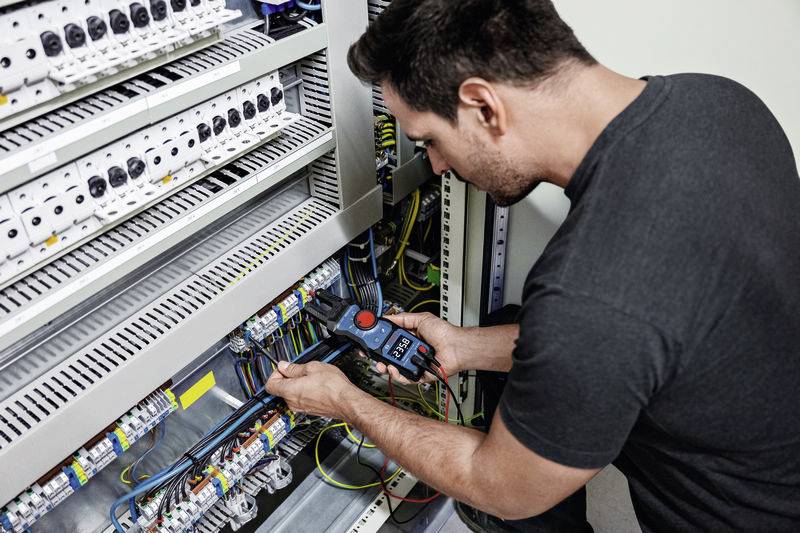 Un homme répare un armoire électrique. Il tient un appareil de mesure et vérifie les connexions de câbles.