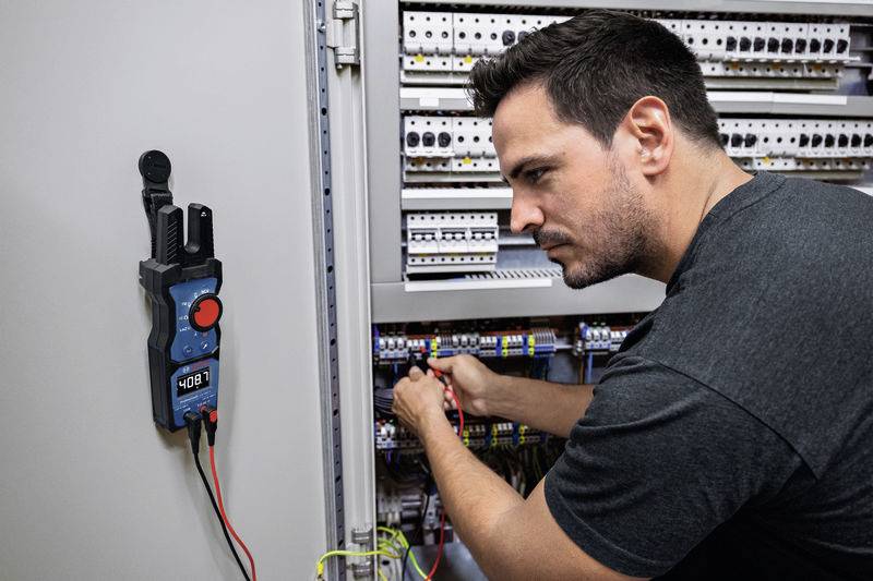 Un homme travaille attentivement sur un tableau électrique ouvert ; un multimètre affiche '408'. L'accent est mis sur une maintenance électrique précise.