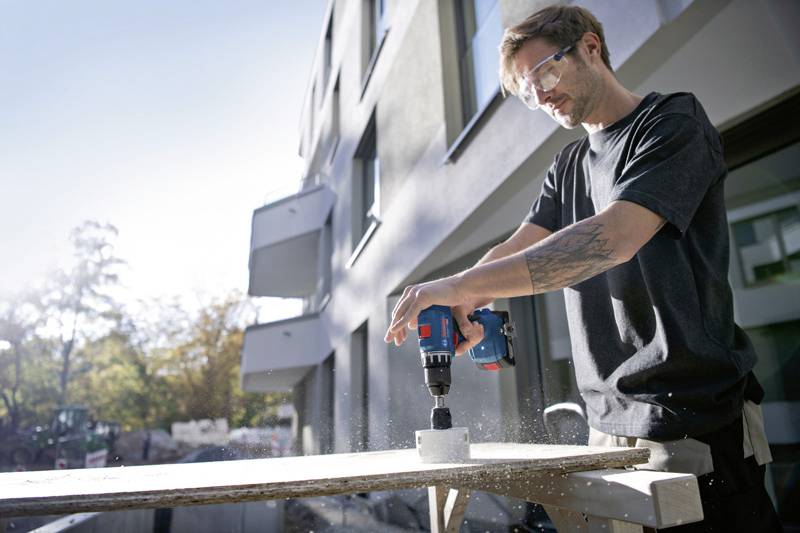 Un homme fore un trou dans une planche en bois avec une perceuse électrique devant un immeuble moderne.