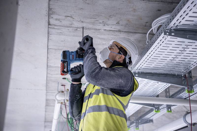 Un ouvrier portant un casque de chantier et un gilet de sécurité fore un plafond. Des câbles et des tuyaux sont visibles. Il porte un masque et des gants de travail.