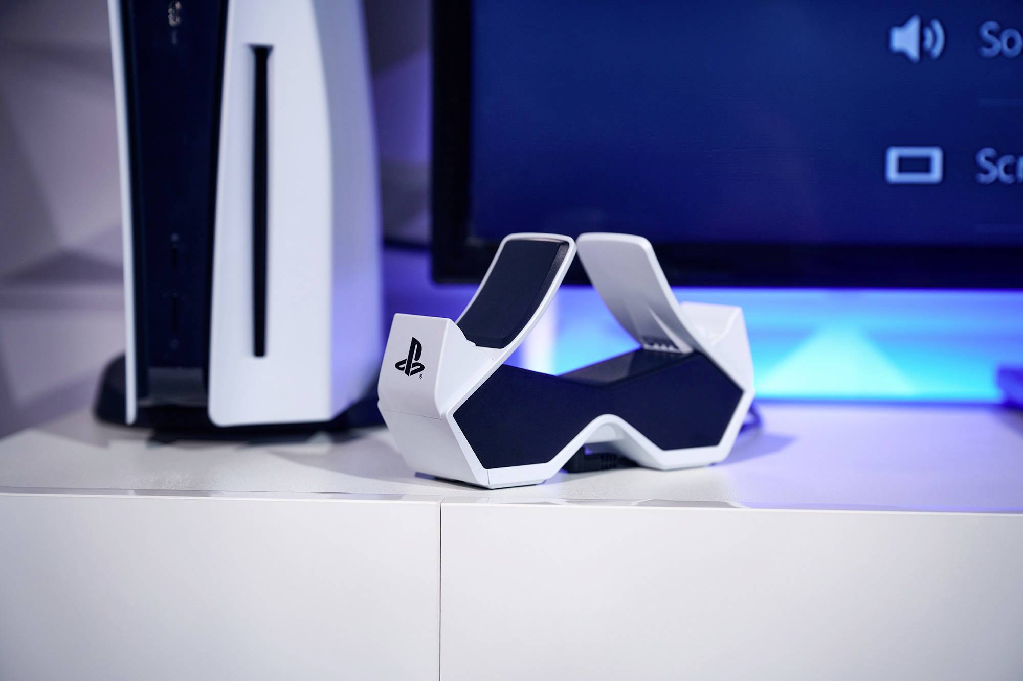 Une console PlayStation 5 et sa station de chargement blanche sur une étagère devant une télévision.