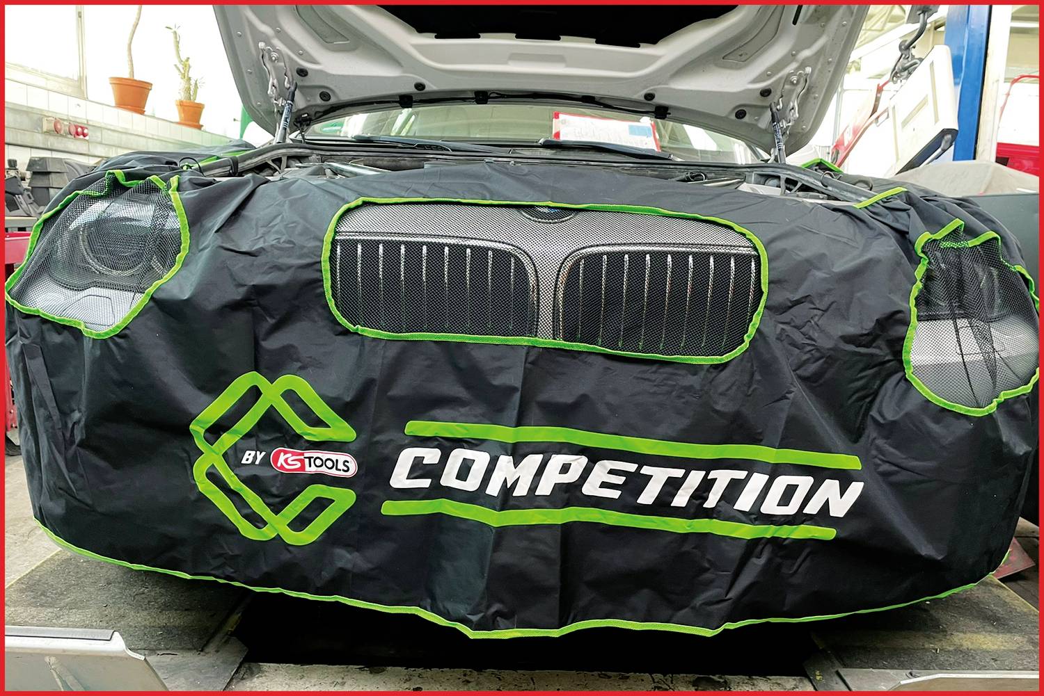Une voiture est garée dans un atelier, recouverte d'une protection noire à l'avant sur laquelle est inscrit « COMPETITION BY KS TOOLS ».