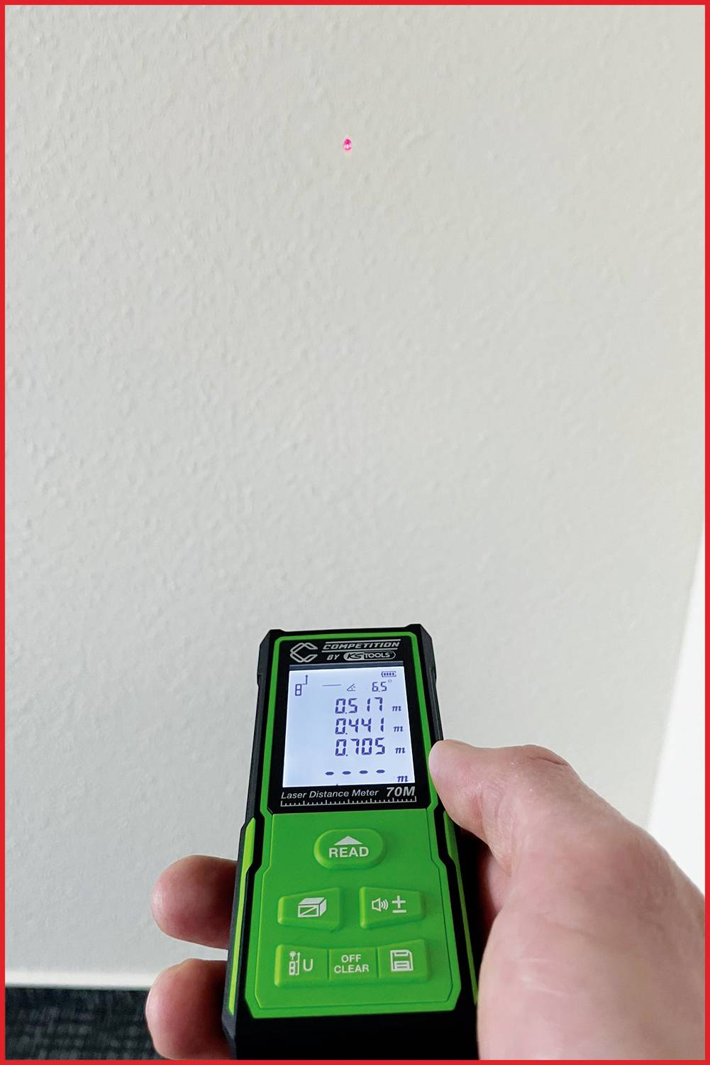 Une main tient un télémètre laser vert. Un point laser rouge est visible sur un mur blanc. L'appareil affiche la mesure de distance.