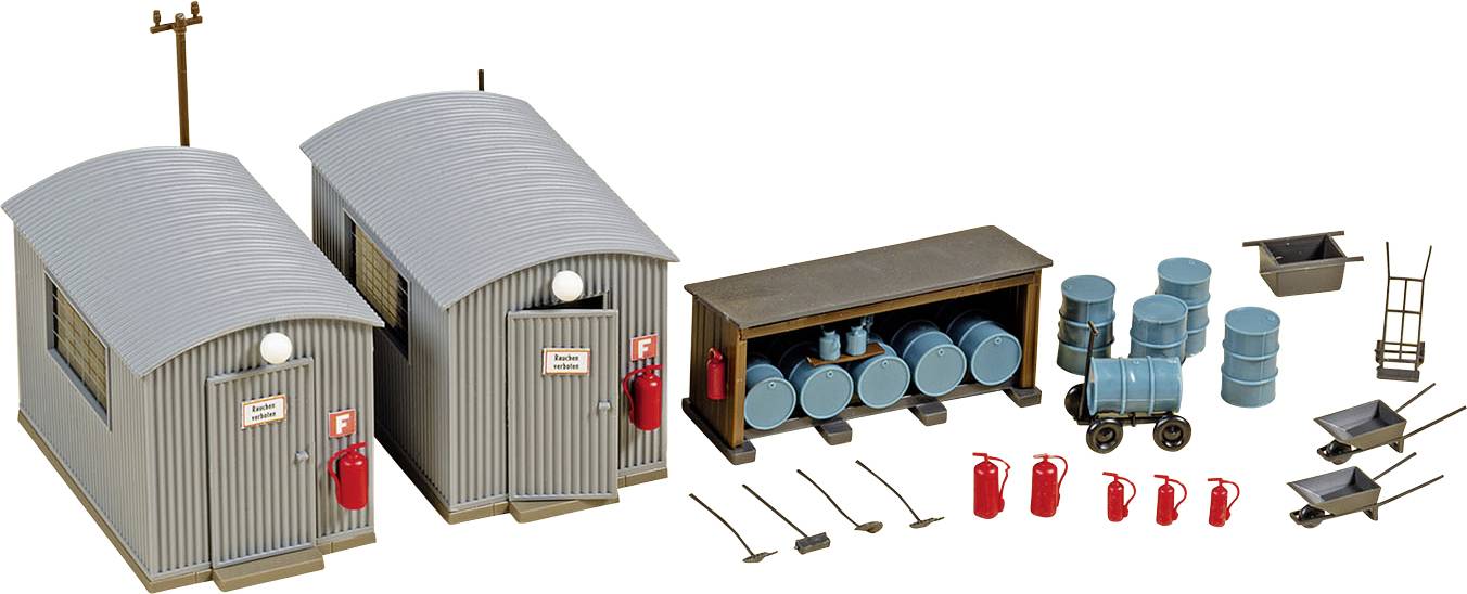 Deux cabanes en tôle ondulée grise avec des extincteurs (marqués 'F'), à côté un espace de stockage ouvert avec des fûts bleus et des extincteurs. Des outils et des brouettes sont également présents.
