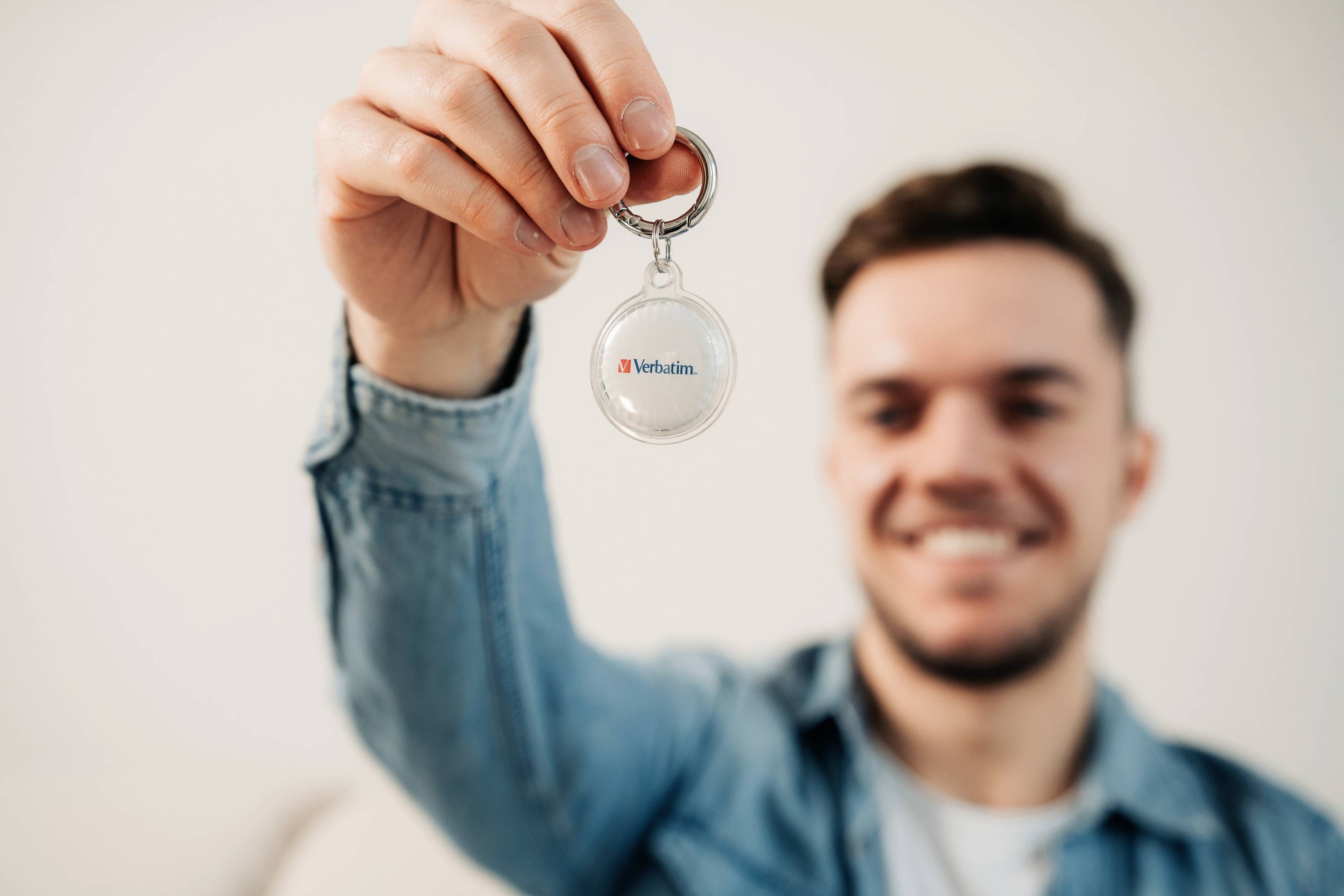Un homme tient en souriant un porte-clés portant l'inscription 'Verbatim' devant l'objectif de la caméra. Il porte une chemise en jean.