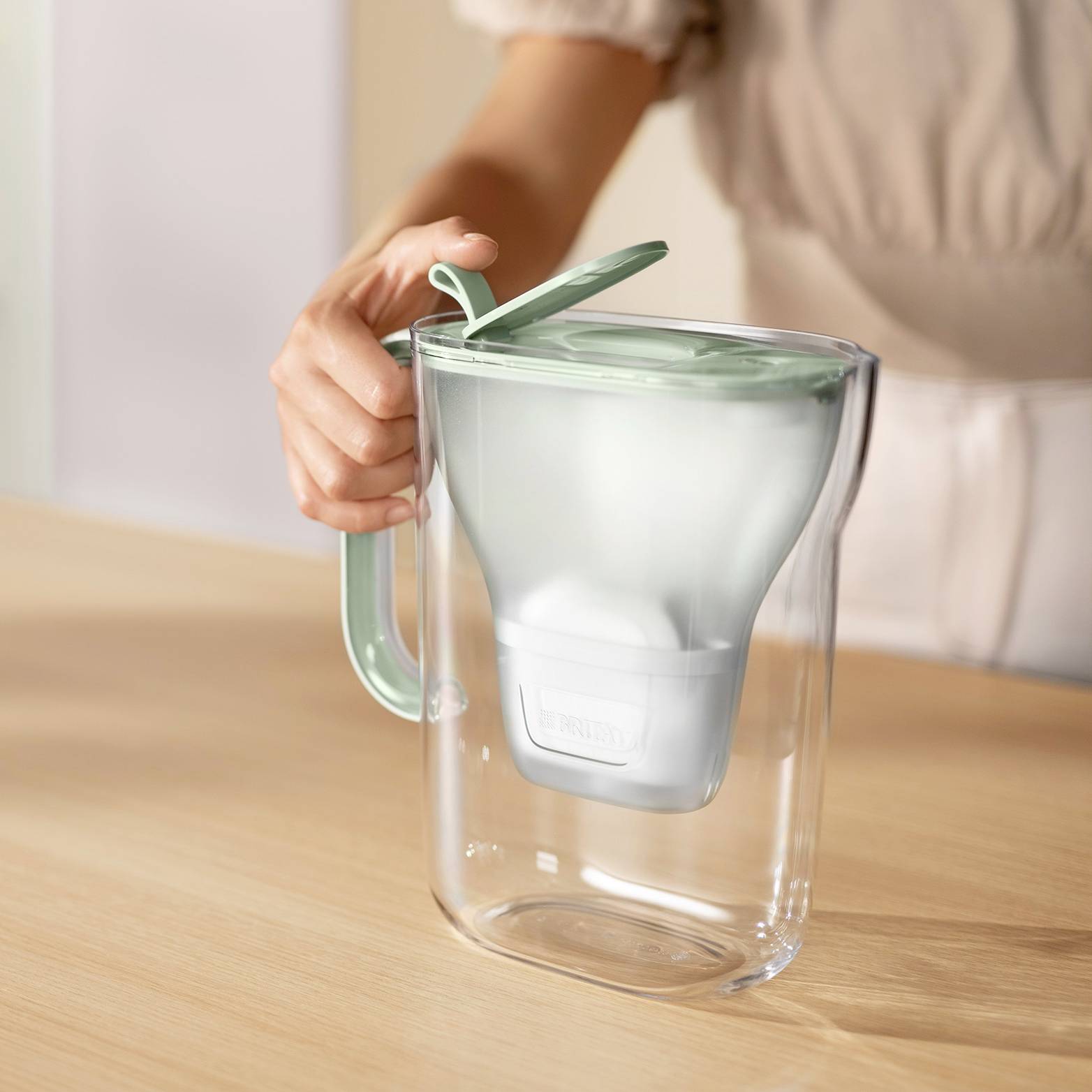 Une personne verse de l'eau d'une carafe filtrante transparente avec un couvercle vert dans une cuisine.