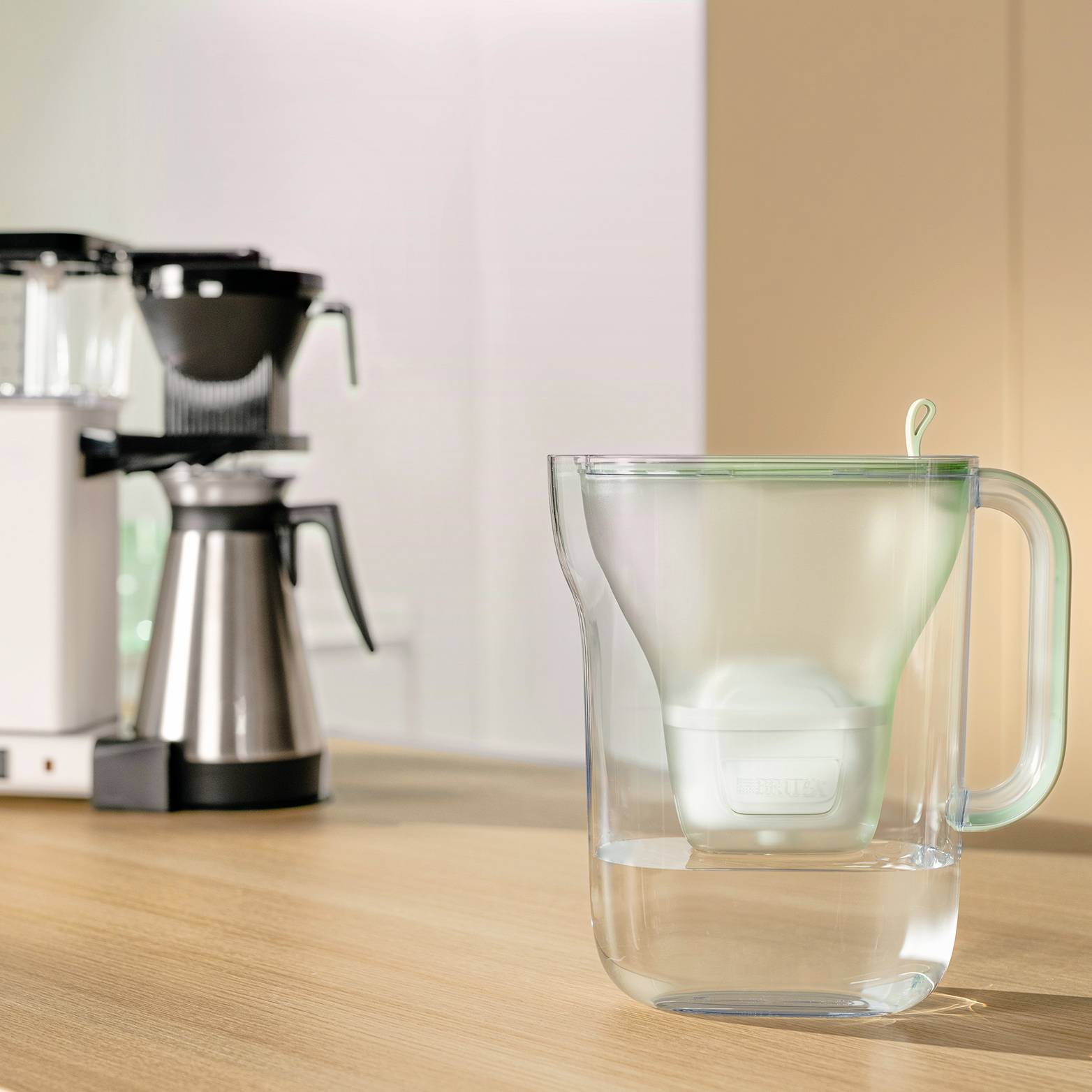 Une carafe d'eau transparente avec un filtre sur une table de cuisine, à côté d'une machine à café et d'autres appareils de cuisine en arrière-plan.
