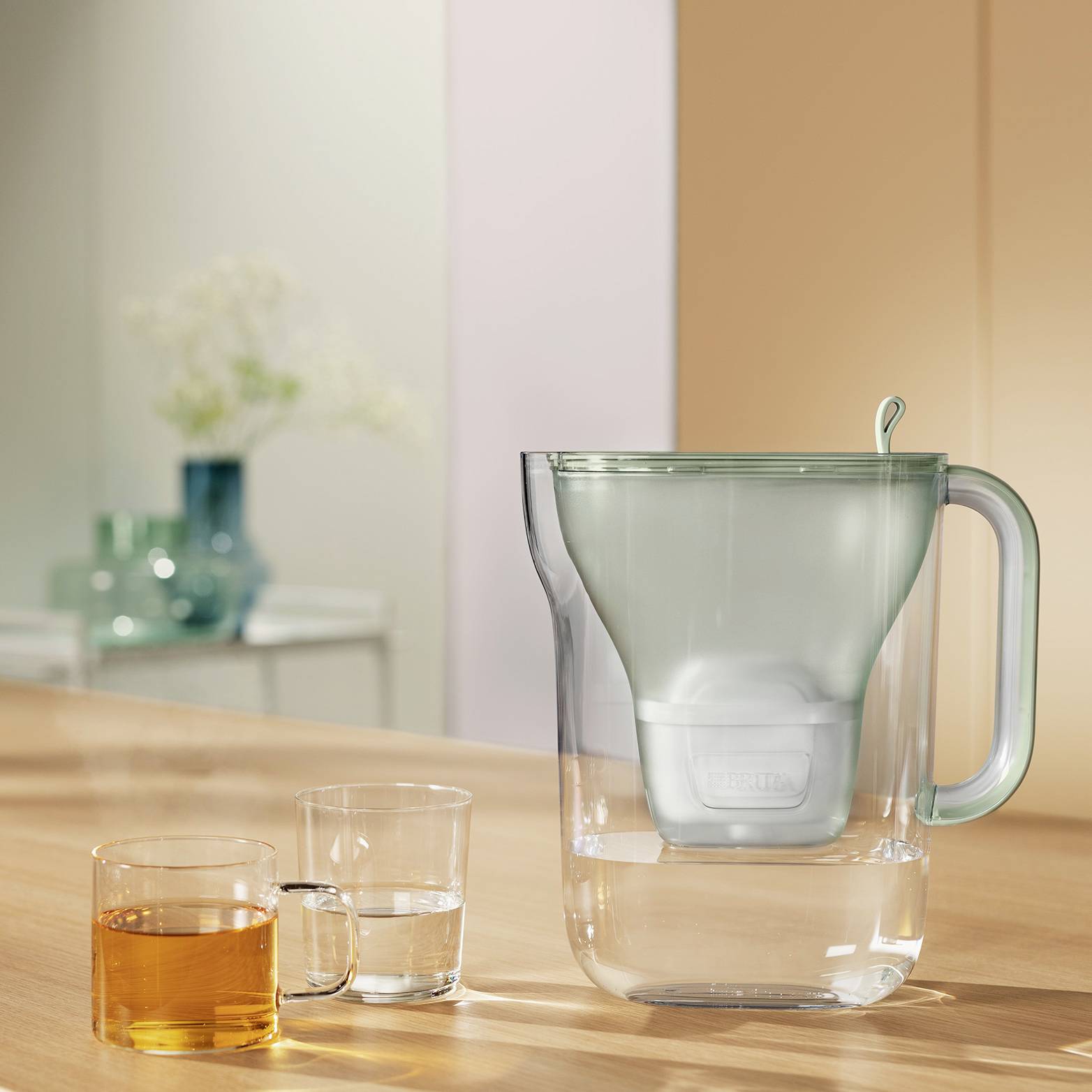 Une carafe d'eau transparente avec un filtre est posée sur une table. À côté se trouvent un verre d'eau et des tasses de thé. En arrière-plan, l'image est floue.