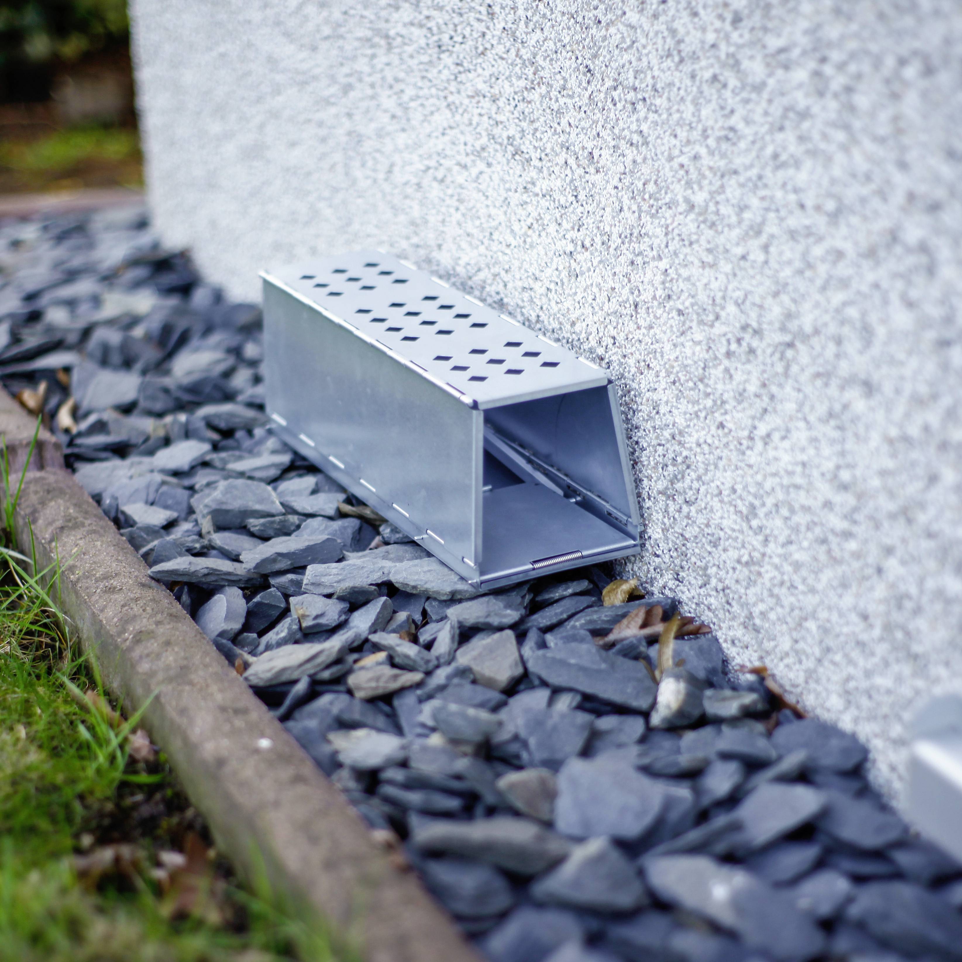 Un chenal de câbles gris court le long de la base d'un mur clair, entouré de pierres gris foncé, dans un espace de jardin.