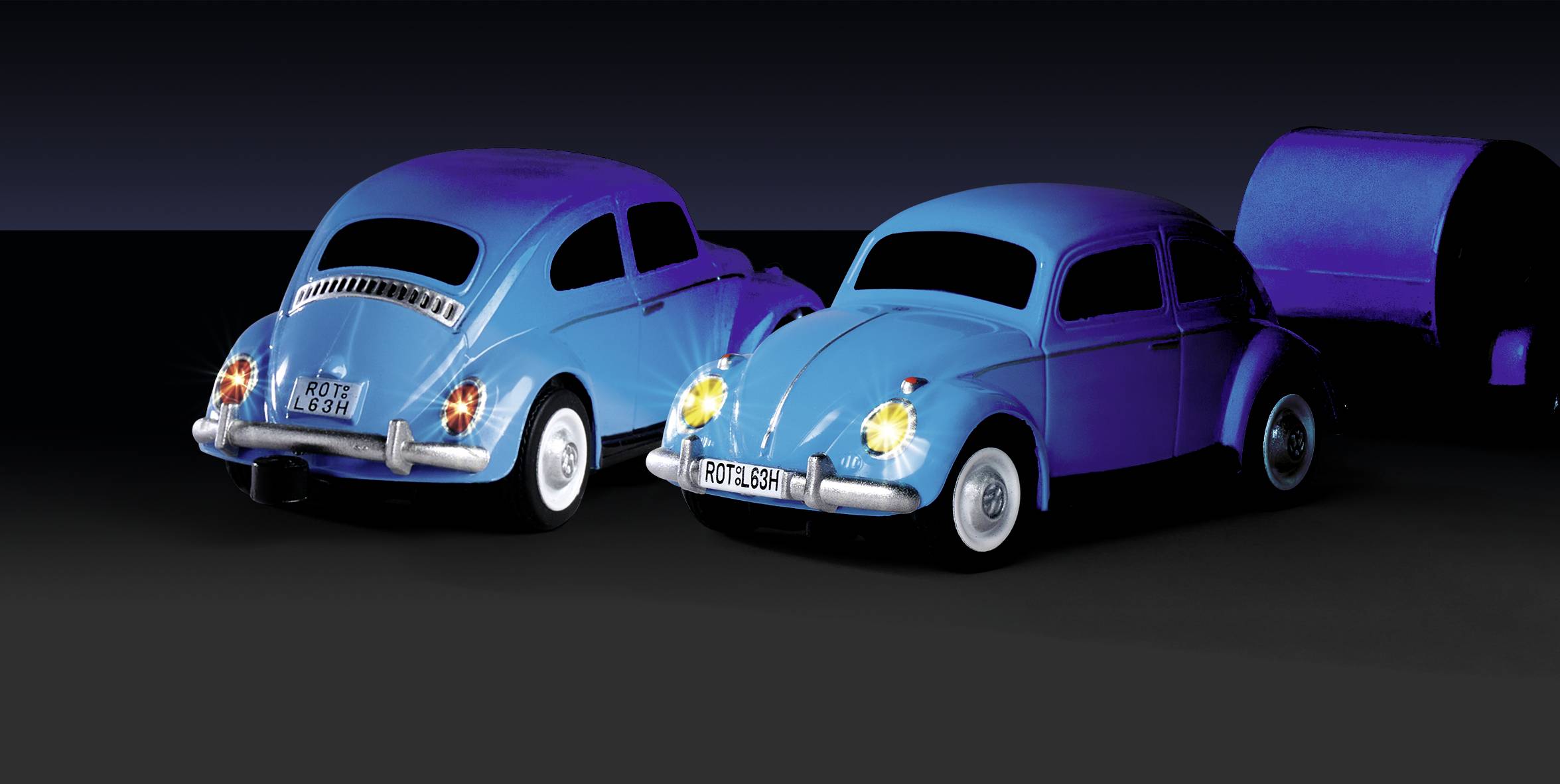 Deux petites voitures jouets bleues ressemblant à un Coccinelle de VW, placées côte à côte. Un remorque est visible en arrière-plan.