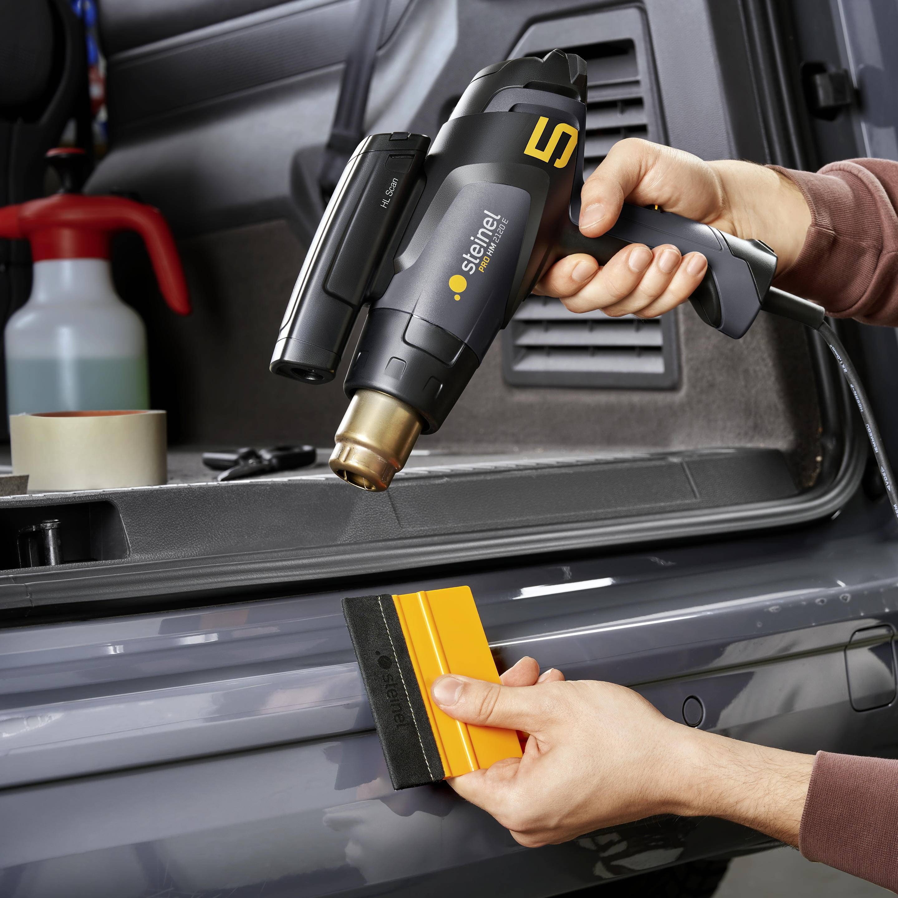 Deux mains utilisent un sèche-cheveux à air chaud pour appliquer un film sur un vernis automobile. Outils et liquide en arrière-plan.