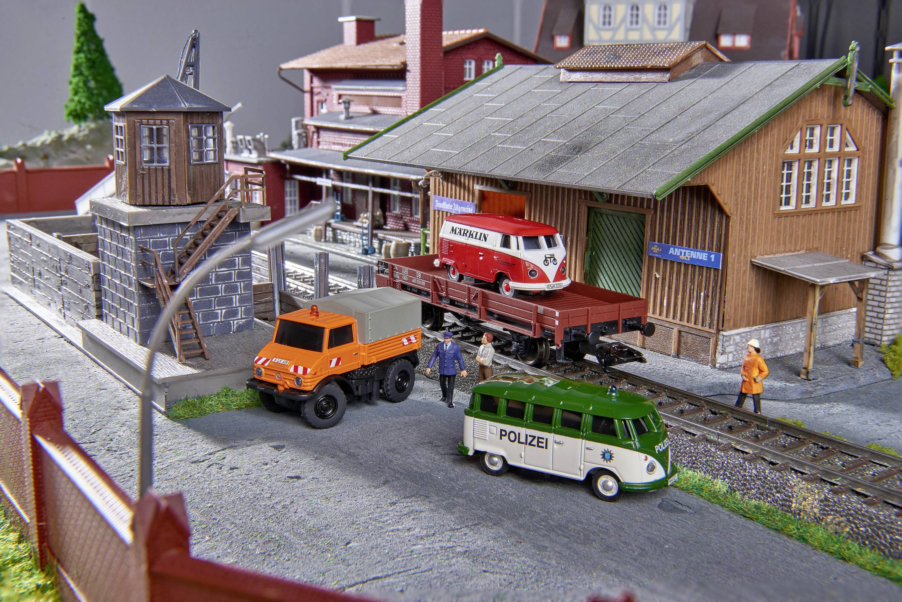 Scène de train miniature avec plusieurs véhicules : Un camion orange, une ambulance et un fourgon de police vert près d'un entrepôt de marchandises. Deux personnages sont debout en tant que travailleurs sur le site.