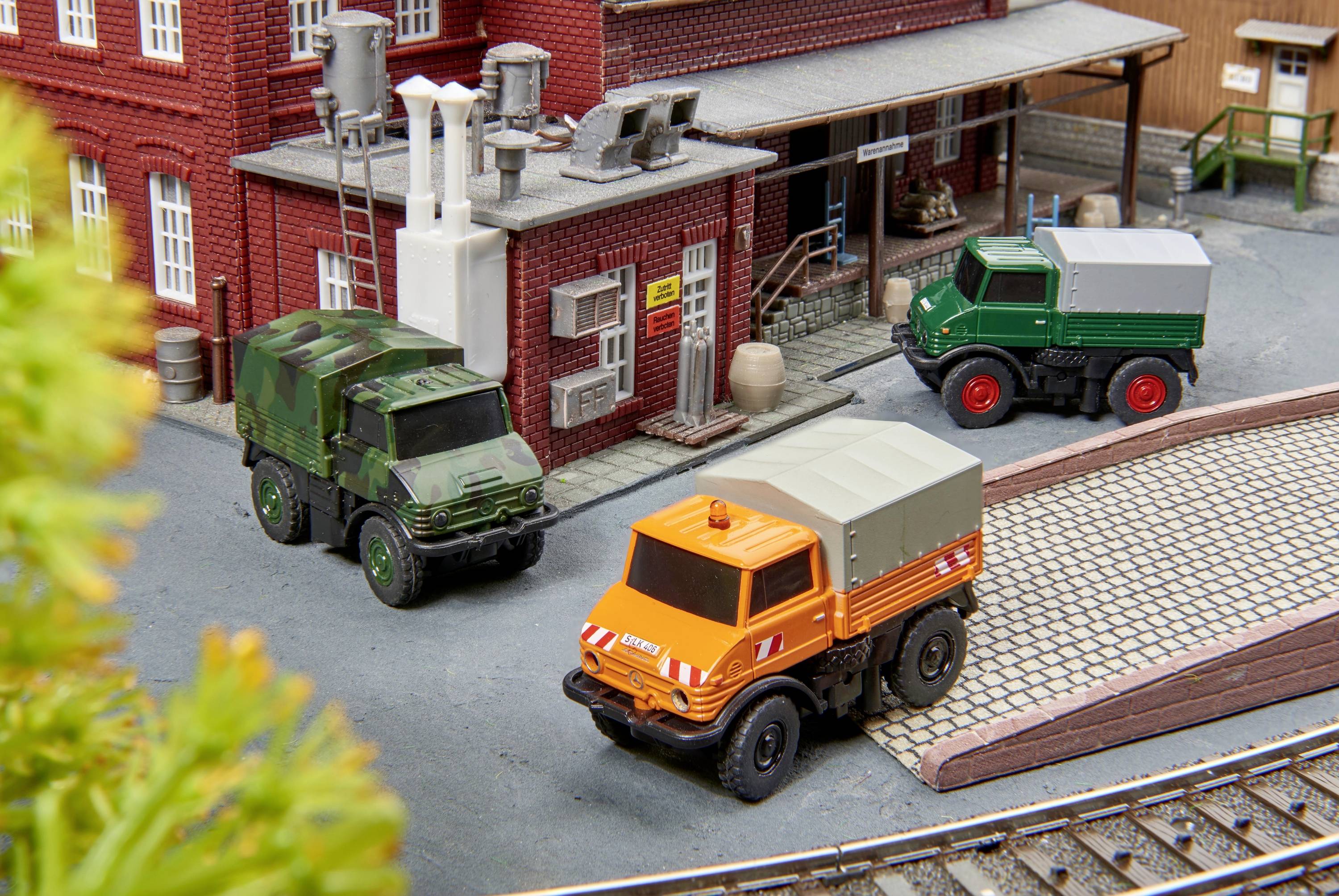 Trois camions-modèles sont garés devant un bâtiment dans une scène miniature. L'un est orange, un autre est peint en vert camouflage, et le troisième est vert.