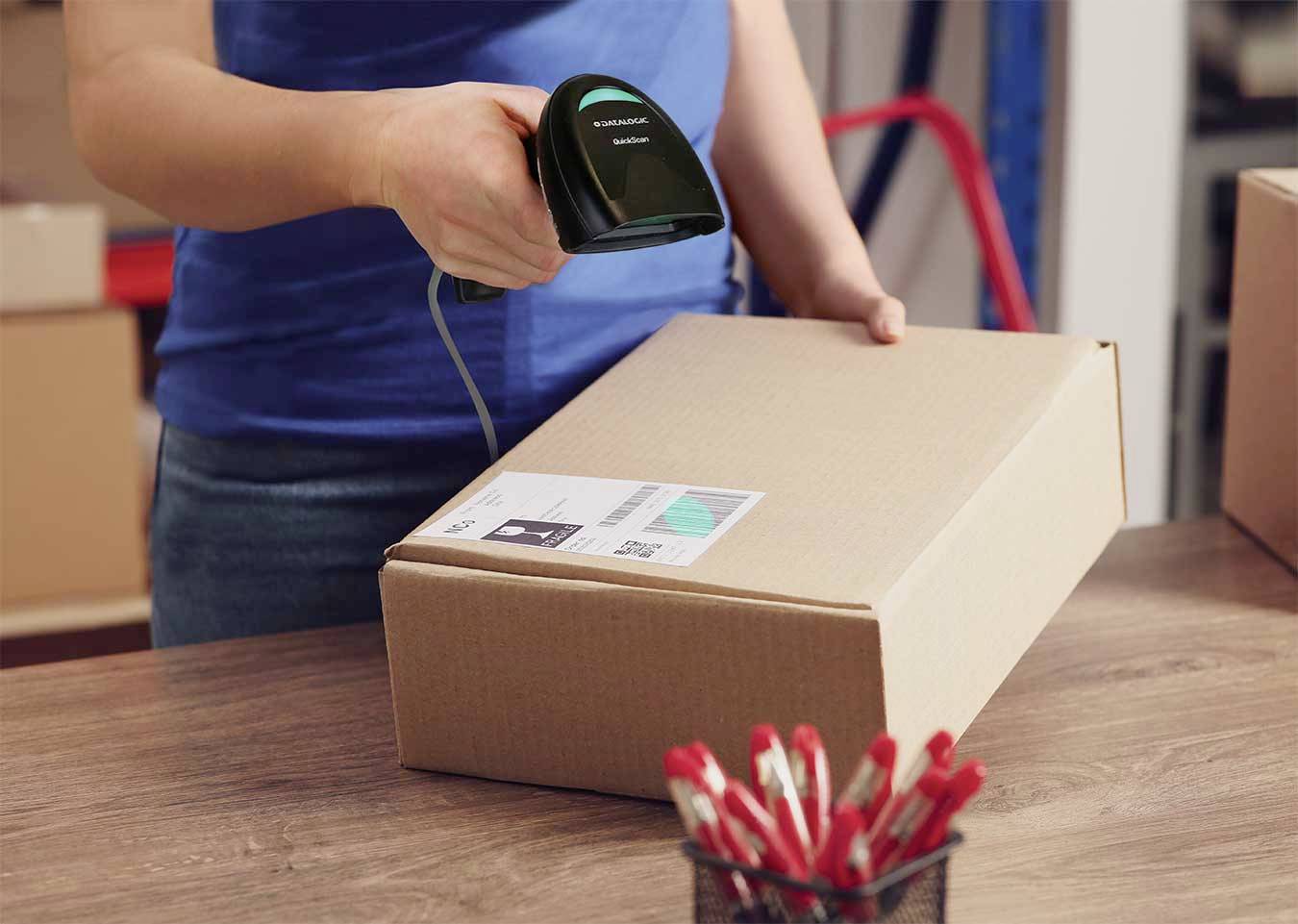 Une personne scanne une étiquette d'expédition sur un carton à l'aide d'un scanner portable sur une table. Au premier plan, un récipient contenant des stylos rouges.