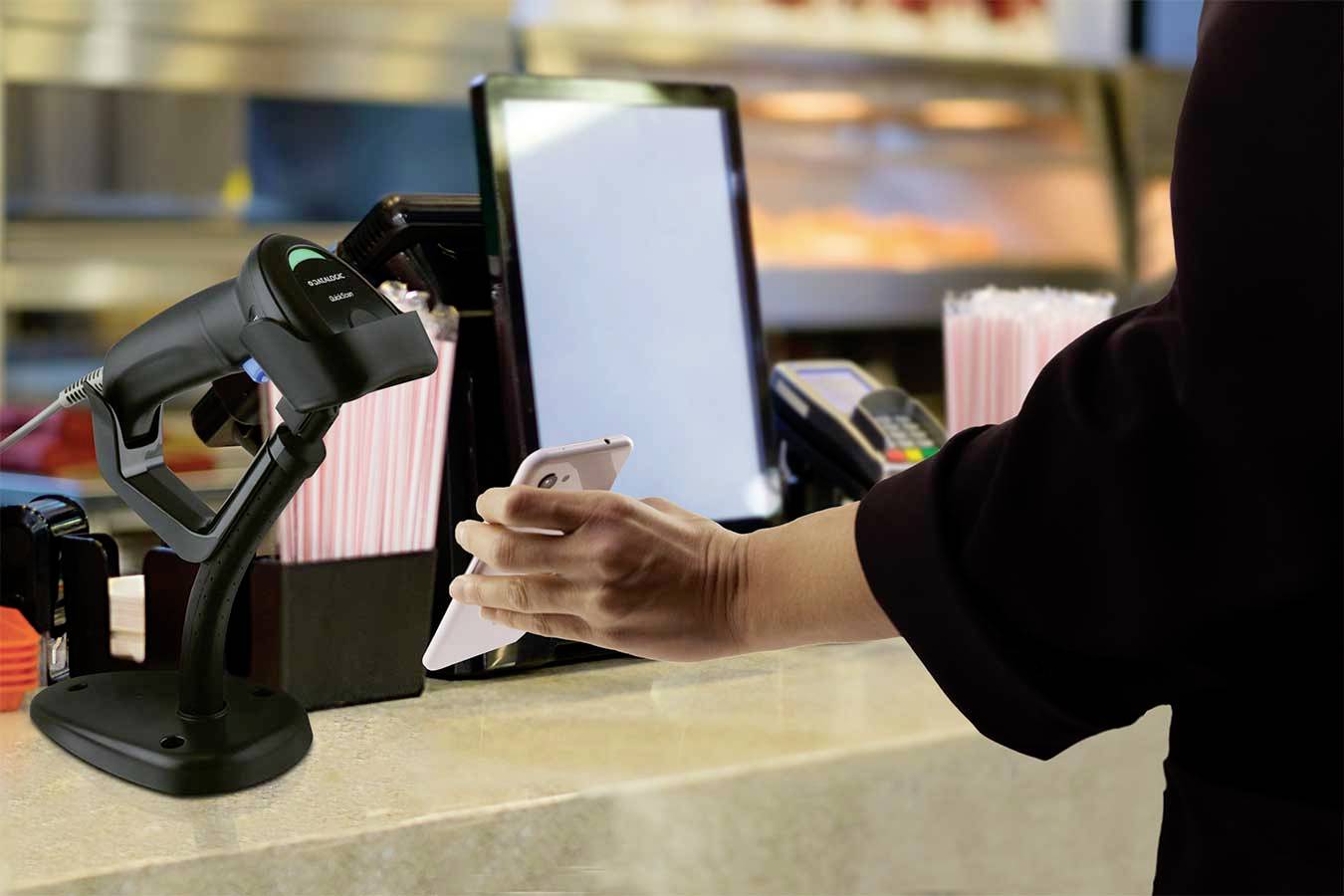 Une personne tient un smartphone près d'un scanner situé sur un comptoir, à côté d'un lecteur de cartes, dans un environnement commercial.