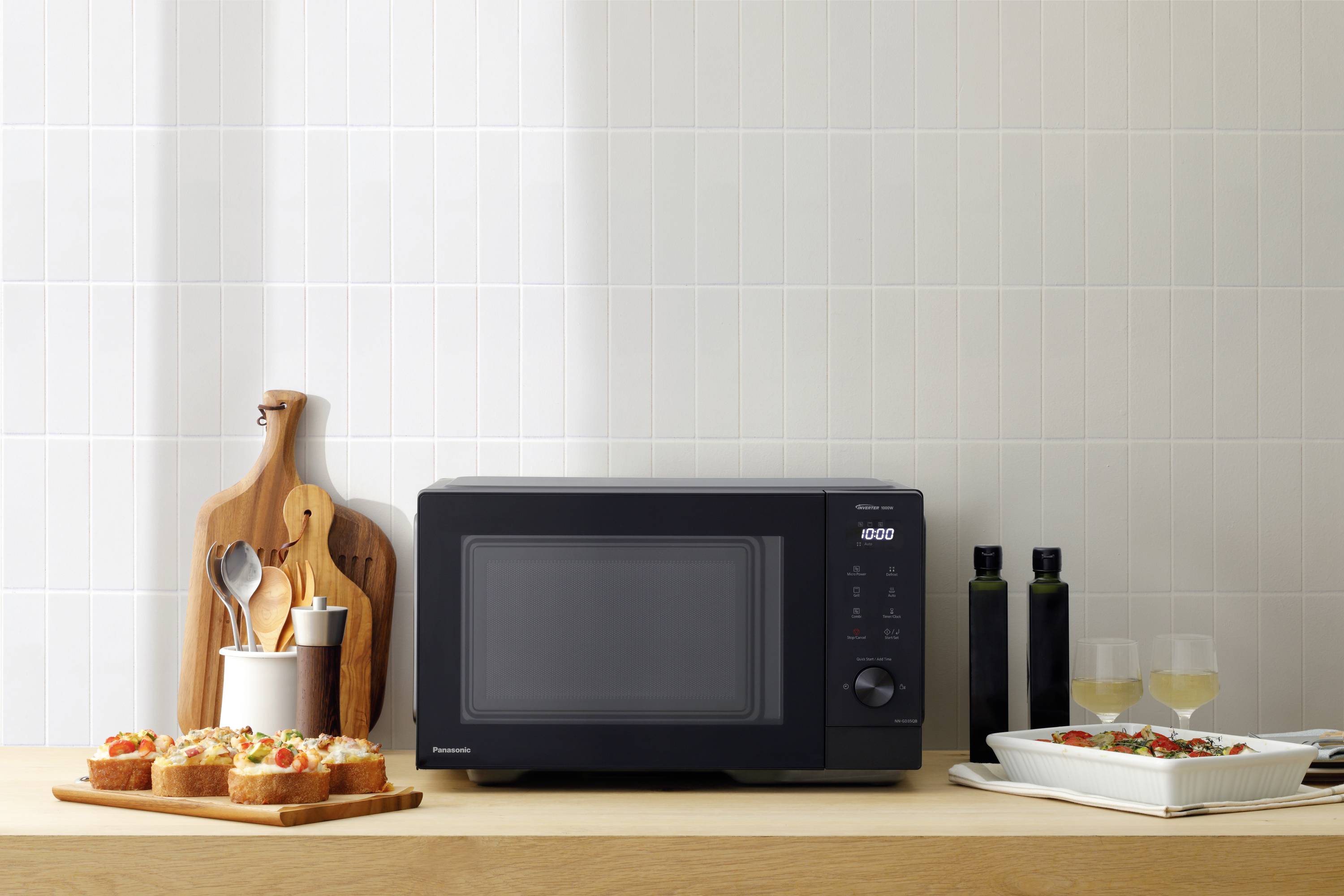 Un micro-ondes noir est posé sur une planche en bois à côté de sandwichs garnis, de deux verres contenant un liquide blanc et d'un plat à gratin blanc.