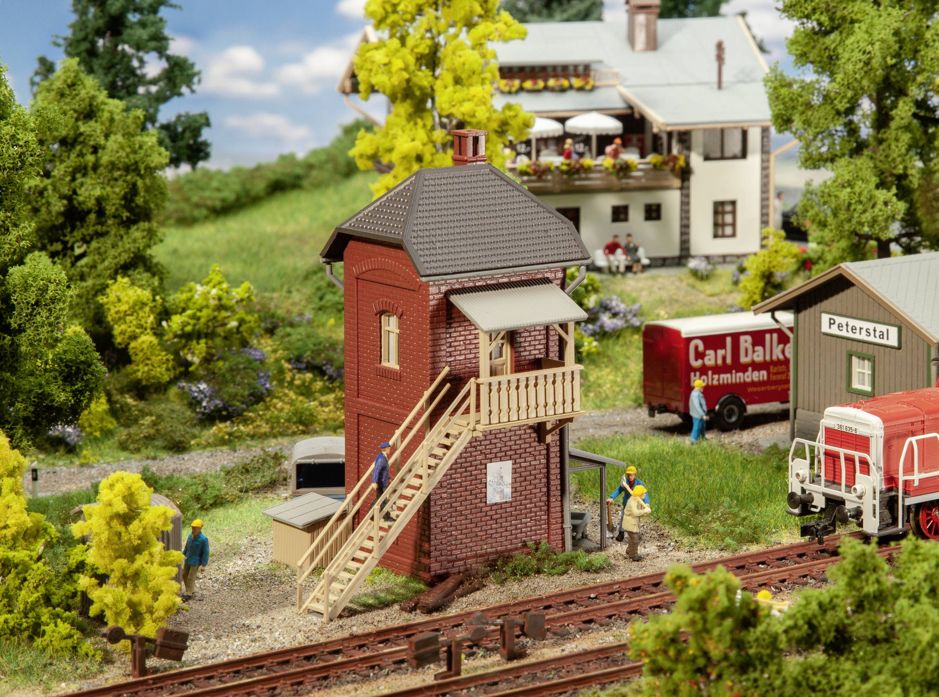 Miniature d'un réseau ferroviaire avec une gare et une locomotive. Des personnes animent la scène. Arrière-plan rural avec des arbres et des maisons.