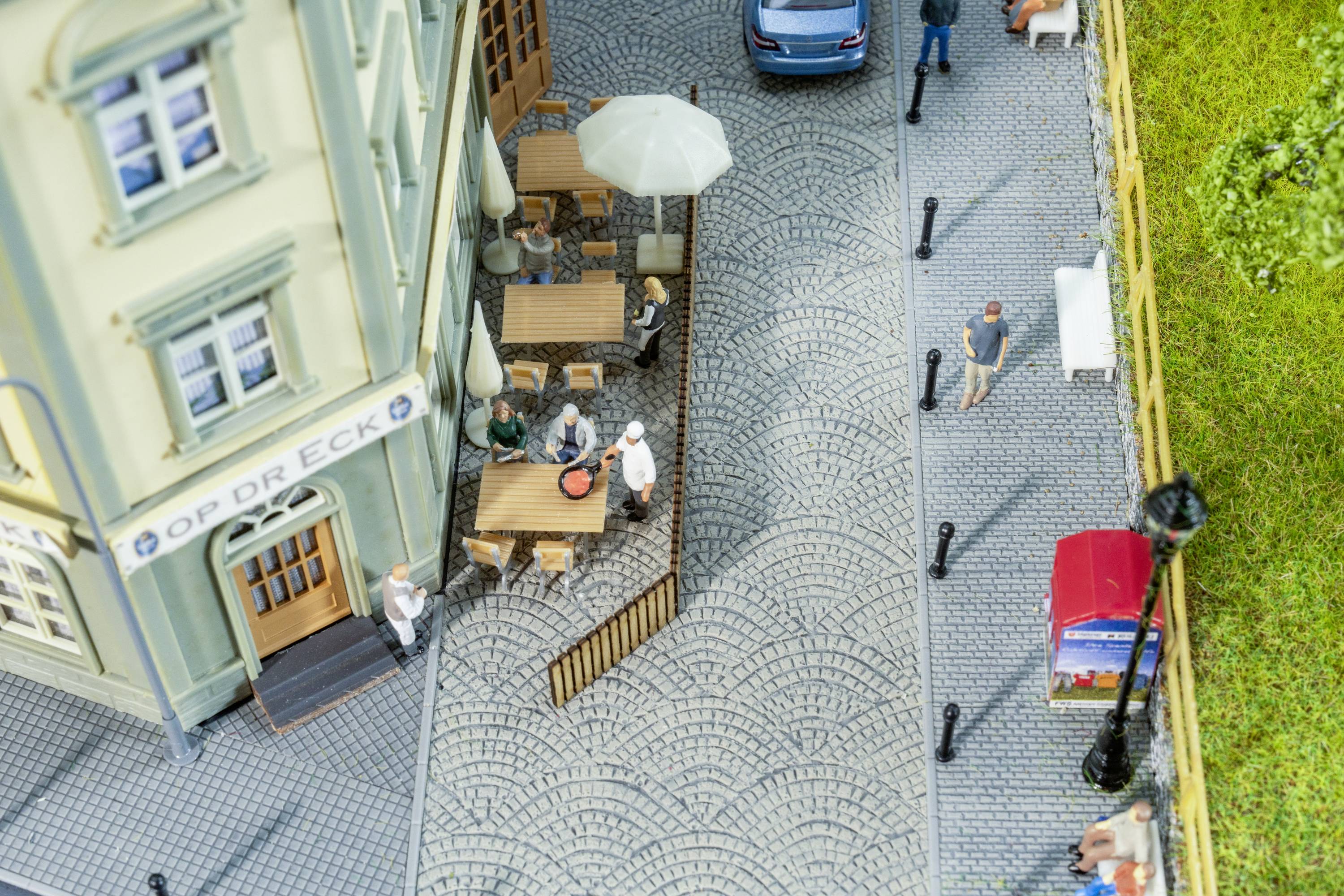 Modèle miniature d'un trottoir avec un café. Trois tables, des clients mangent et boivent. Une voiture bleue dans la rue, des gens se promènent.