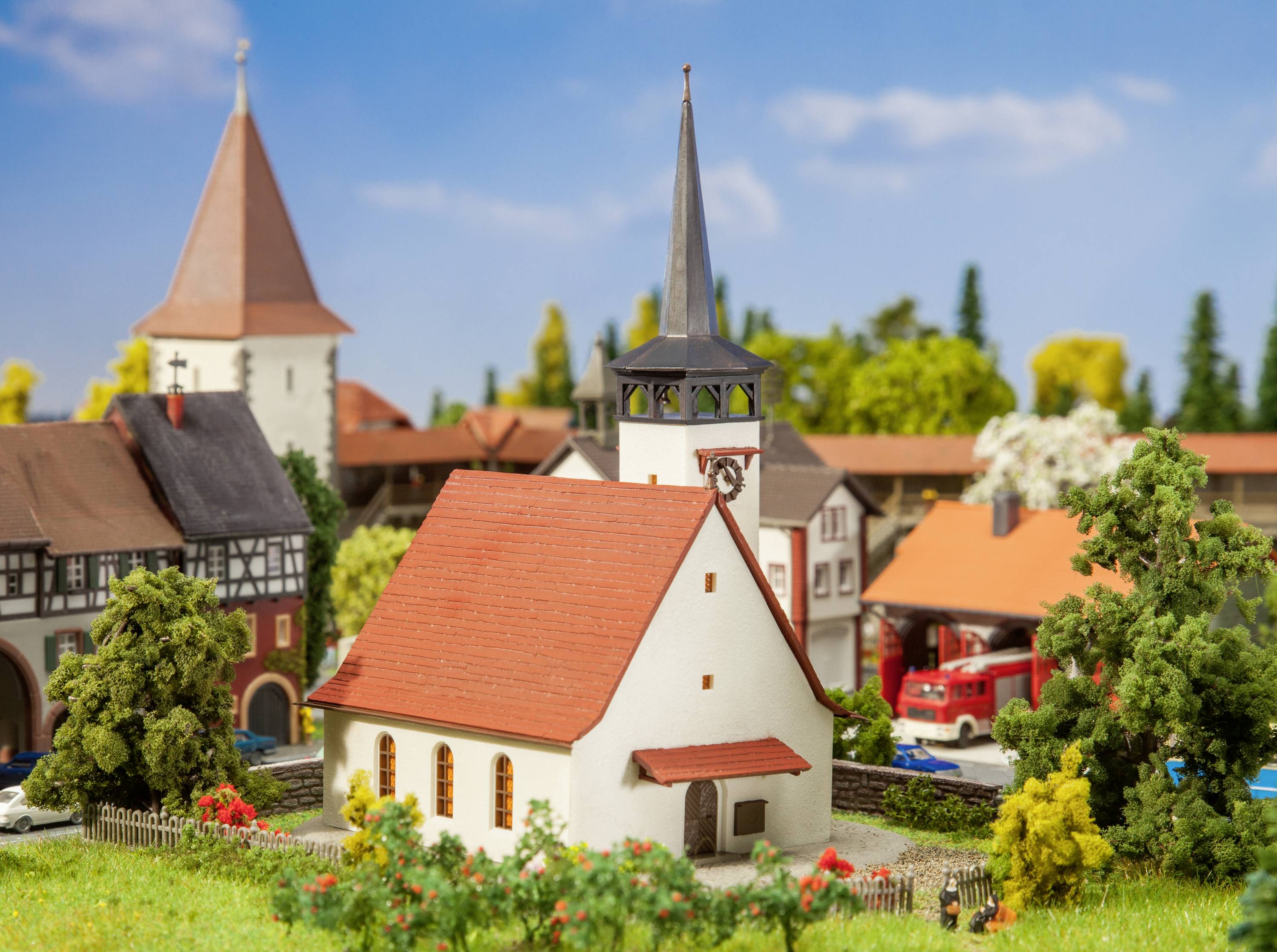 Modèle d'une église de village pittoresque au toit rouge, à côté d'un camion de pompiers, dans un village miniature comprenant plusieurs bâtiments et arbres.