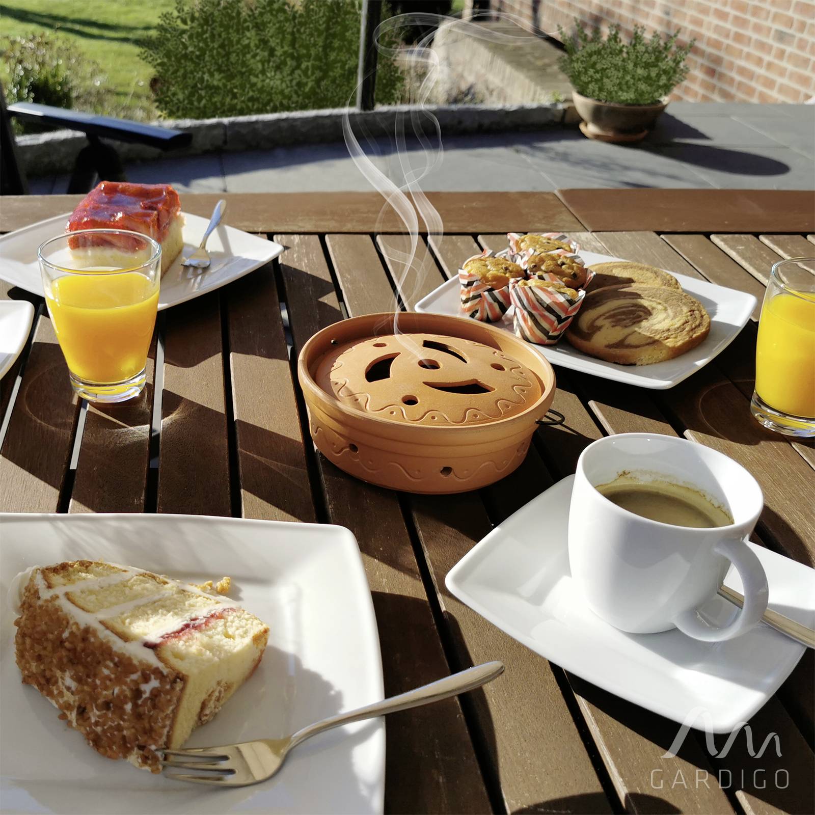 Table du petit-déjeuner en extérieur. Sur une table en bois sont disposés du café, du jus d'orange, des parts de gâteau et des pâtisseries. La lumière du soleil illumine la scène.