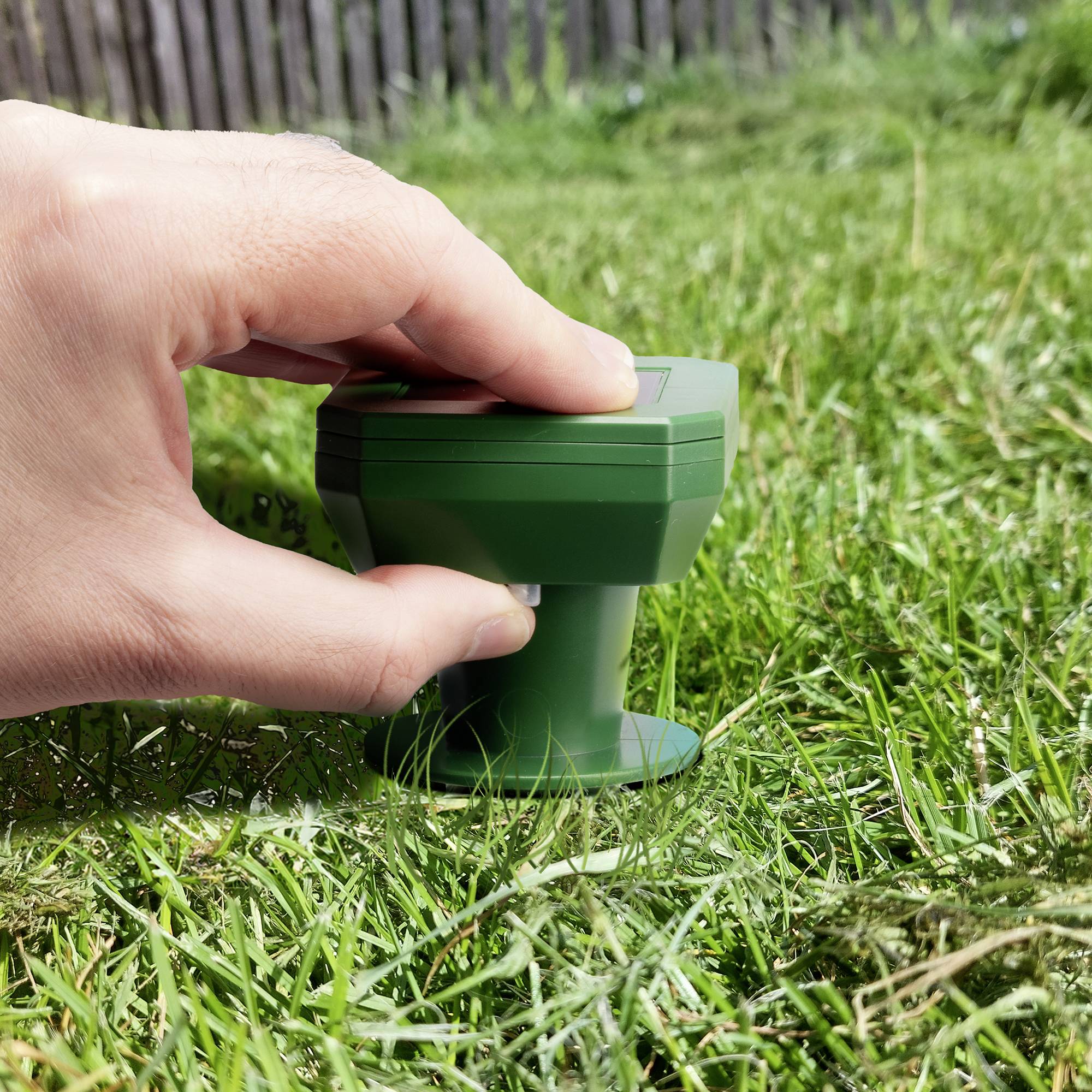 Une main place un arroseur d'herbe vert sur un gazon vert. Une clôture en bois est visible en arrière-plan.