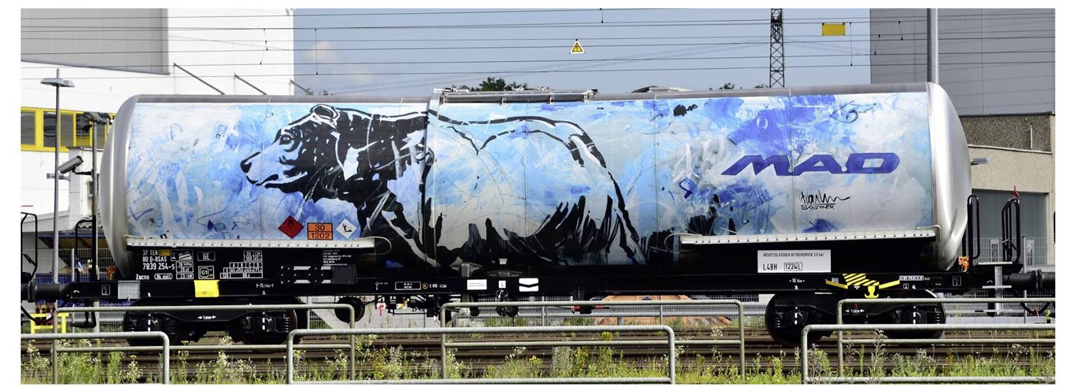 Un wagon-citerne sur un train, peint avec un grand ours noir sur un fond bleu. Texte 'MAD' et marquages de sécurité visibles.