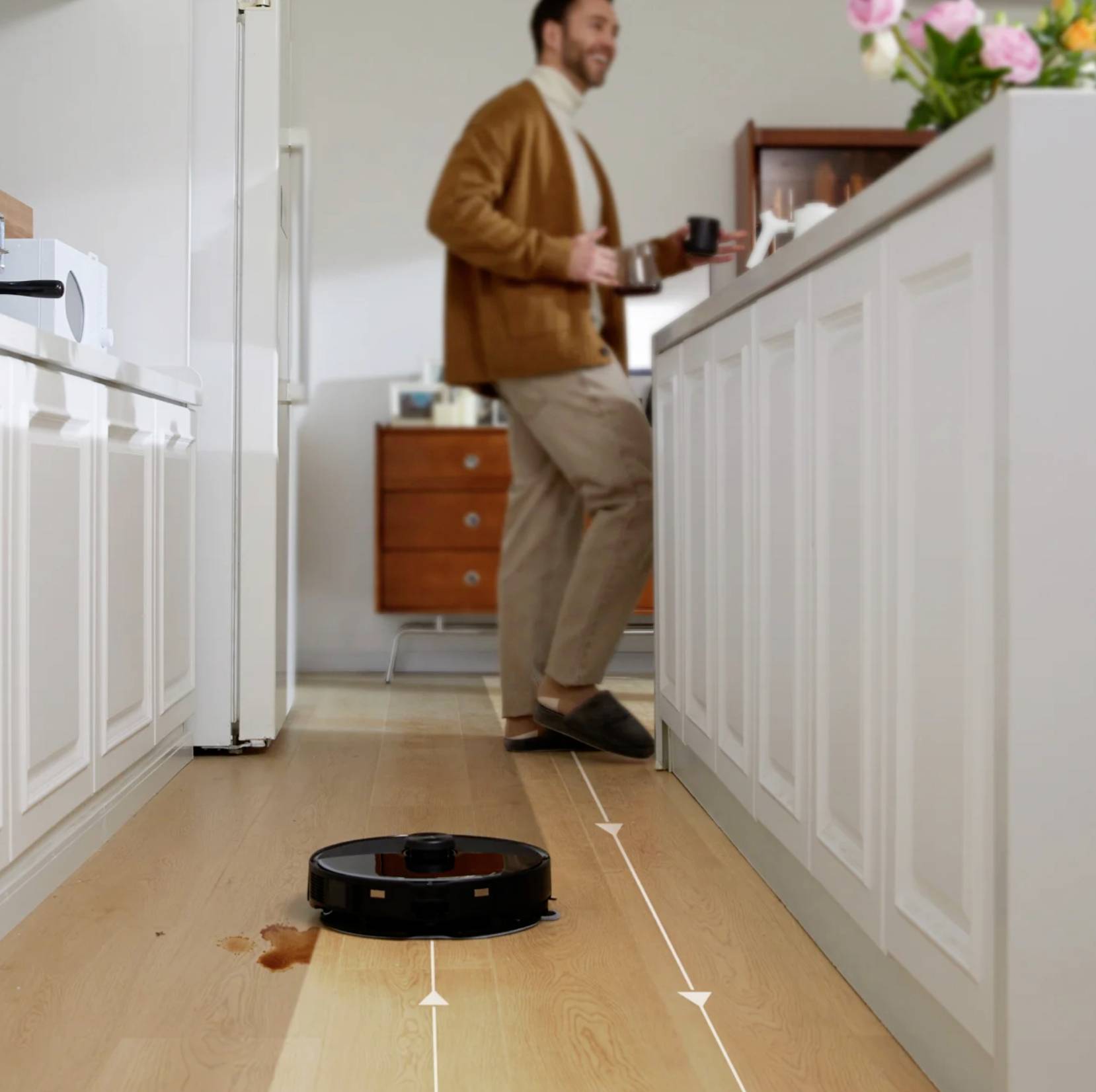 Un homme boit du café dans la cuisine pendant qu'un aspirateur robot nettoie le sol. Au premier plan, le robot est en train de travailler.