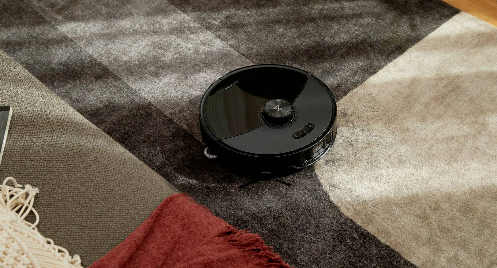Un robot aspirateur noir nettoie un tapis à motifs à côté d'un canapé recouvert de couvertures rouges et beiges.