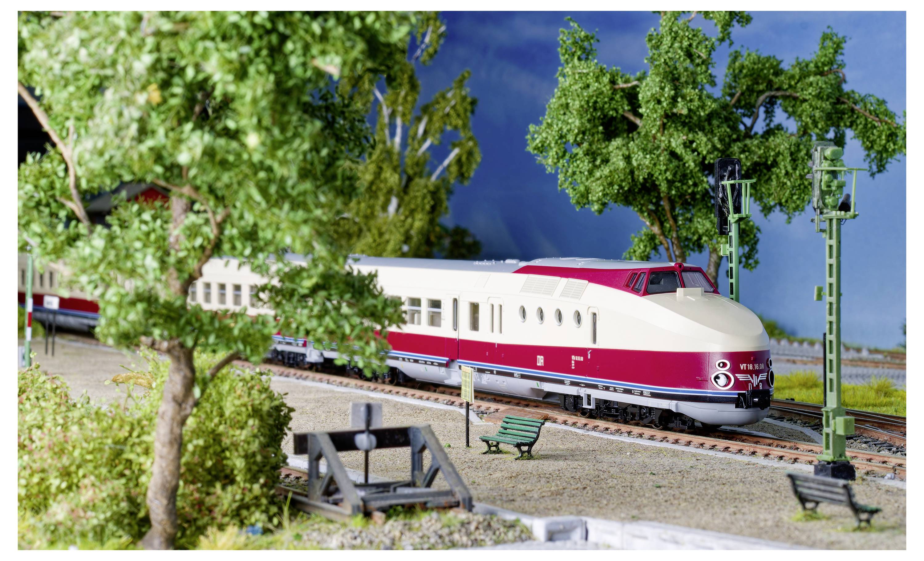 Un train miniature rouge et blanc traverse un paysage détaillé avec des arbres, des bancs et une petite gare.