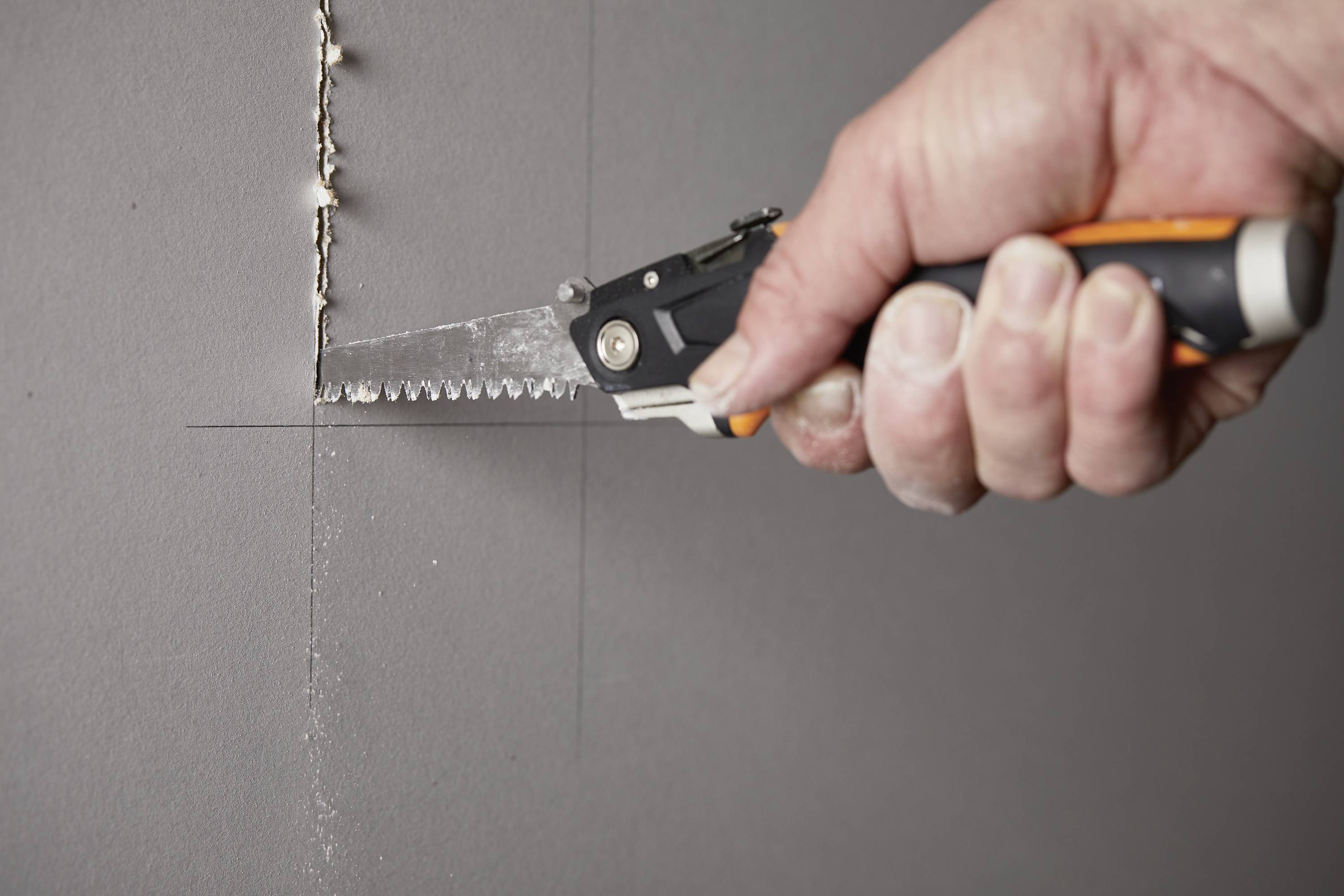 Une main tient une scie et coupe le long d'une ligne sur un mur gris.
