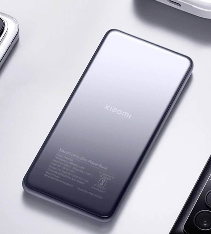 Une batterie externe fine et métallique portant l'étiquette 'Xiaomi' est posée sur une surface claire, entourée de pièces d'autres appareils électroniques.
