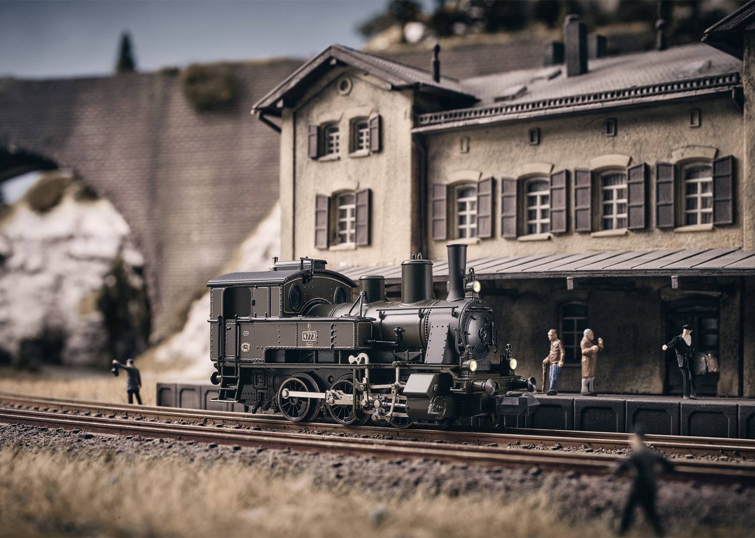 Train miniature avec un train à vapeur historique devant une ancienne gare. Quelques personnages sont sur le quai. L'arrière-plan montre un pont en pierre.