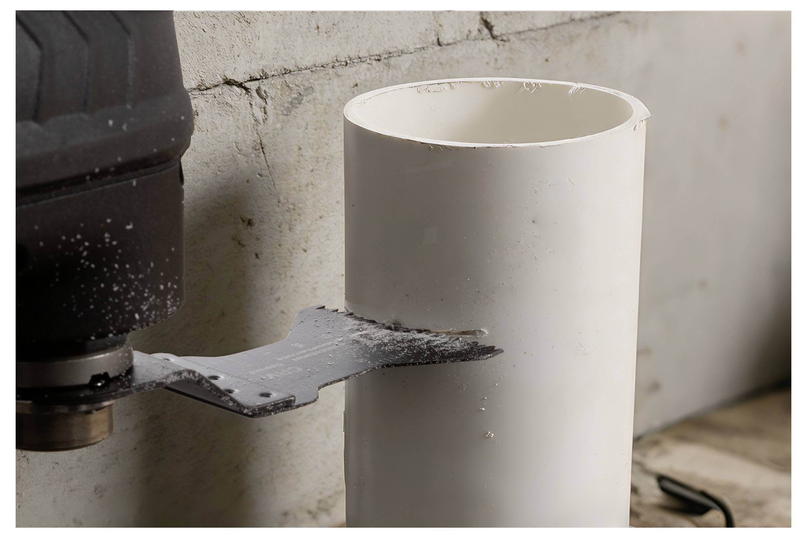 Une lame de scie en train de couper un tuyau vertical blanc en PVC, avec un mur en béton et un outil électrique visibles, illustrant un processus de coupe de tuyau.