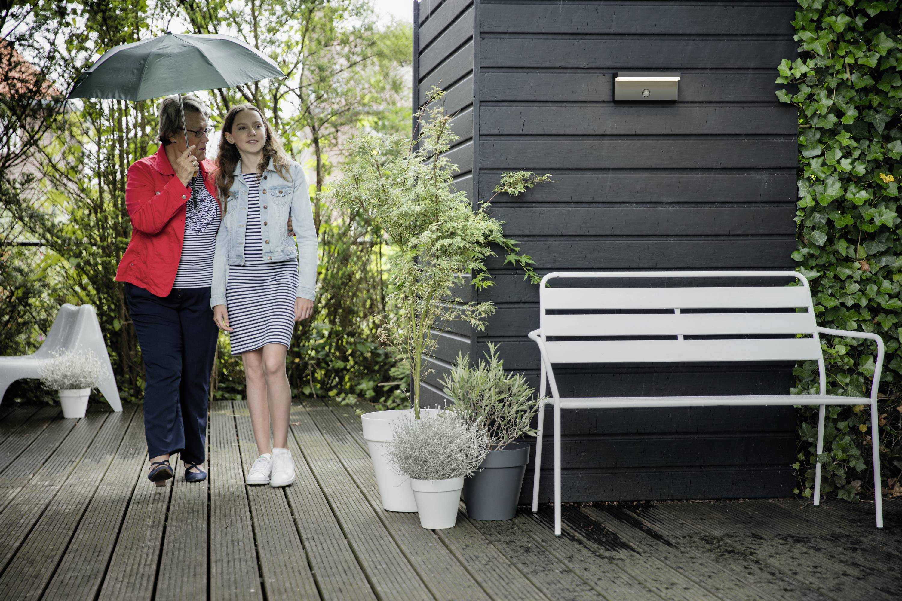 Deux personnes sous un parapluie se tiennent sur une terrasse en bois à côté d'un banc et de plantes. Une personne tient le parapluie au-dessus d'elles deux.