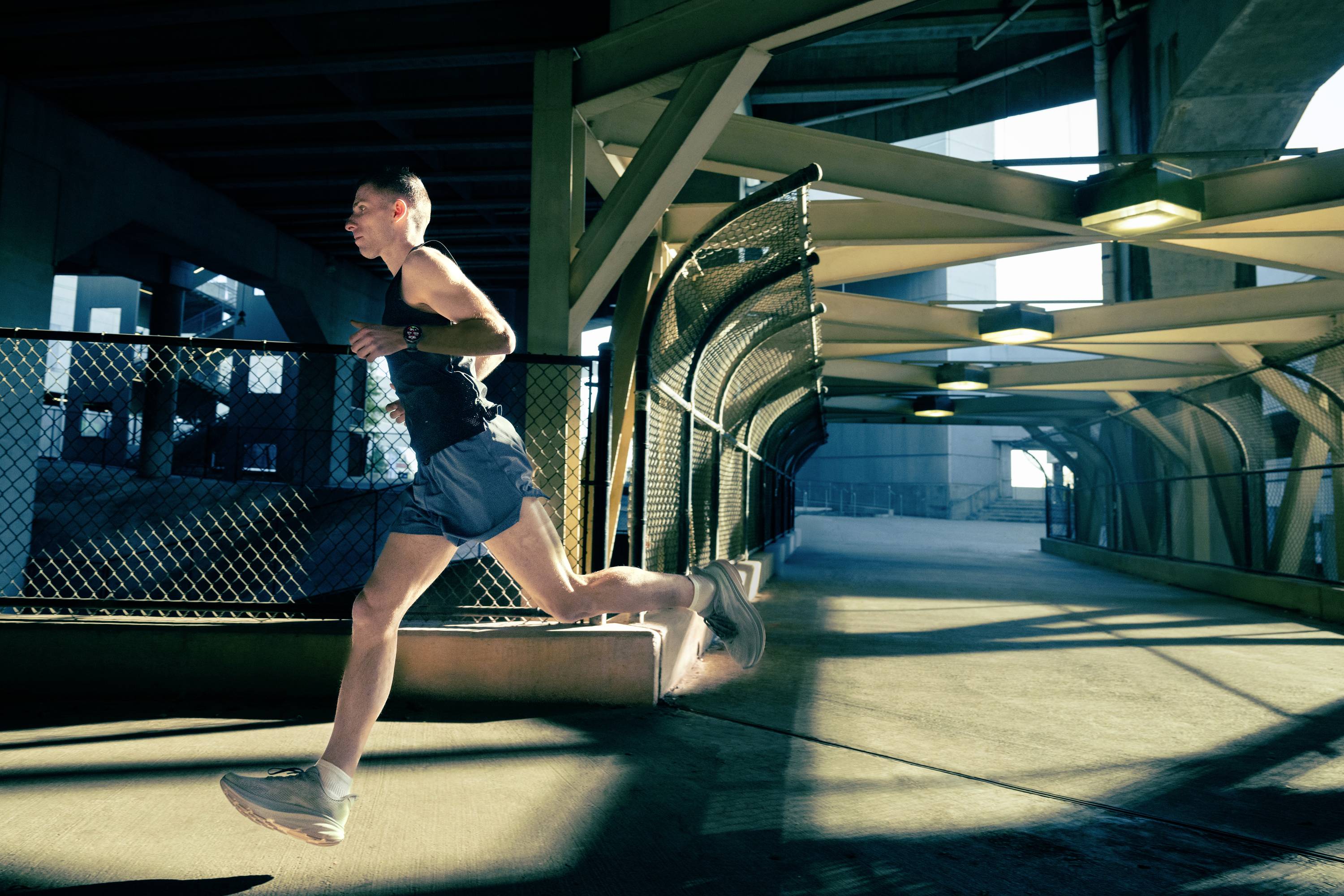 Une personne fait du jogging sur un chemin ombragé sous un pont. La lumière du soleil filtre à travers la structure, créant des jeux d'ombres intéressants.